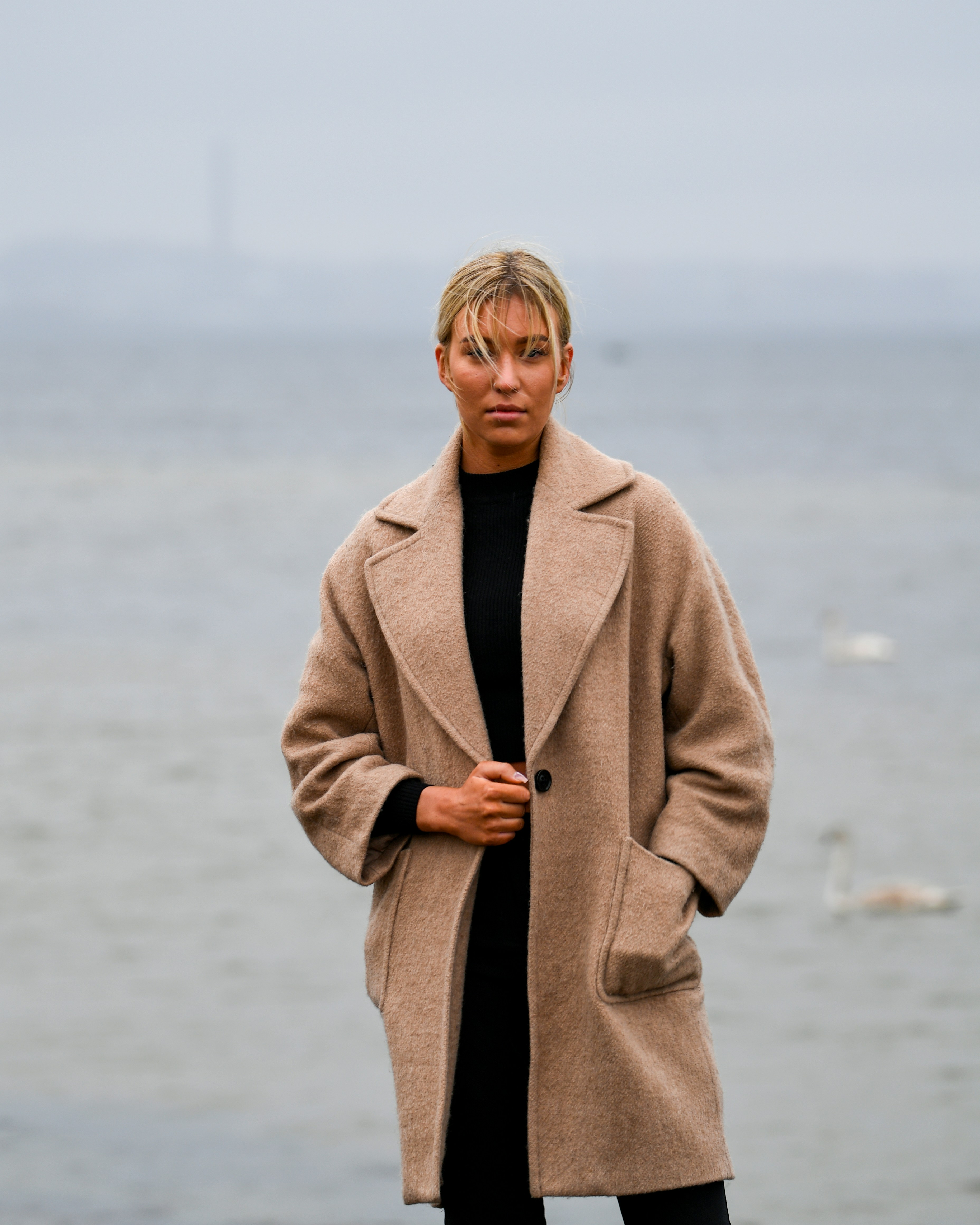 Eine Frau im beigen Mantel steht am Wasser.