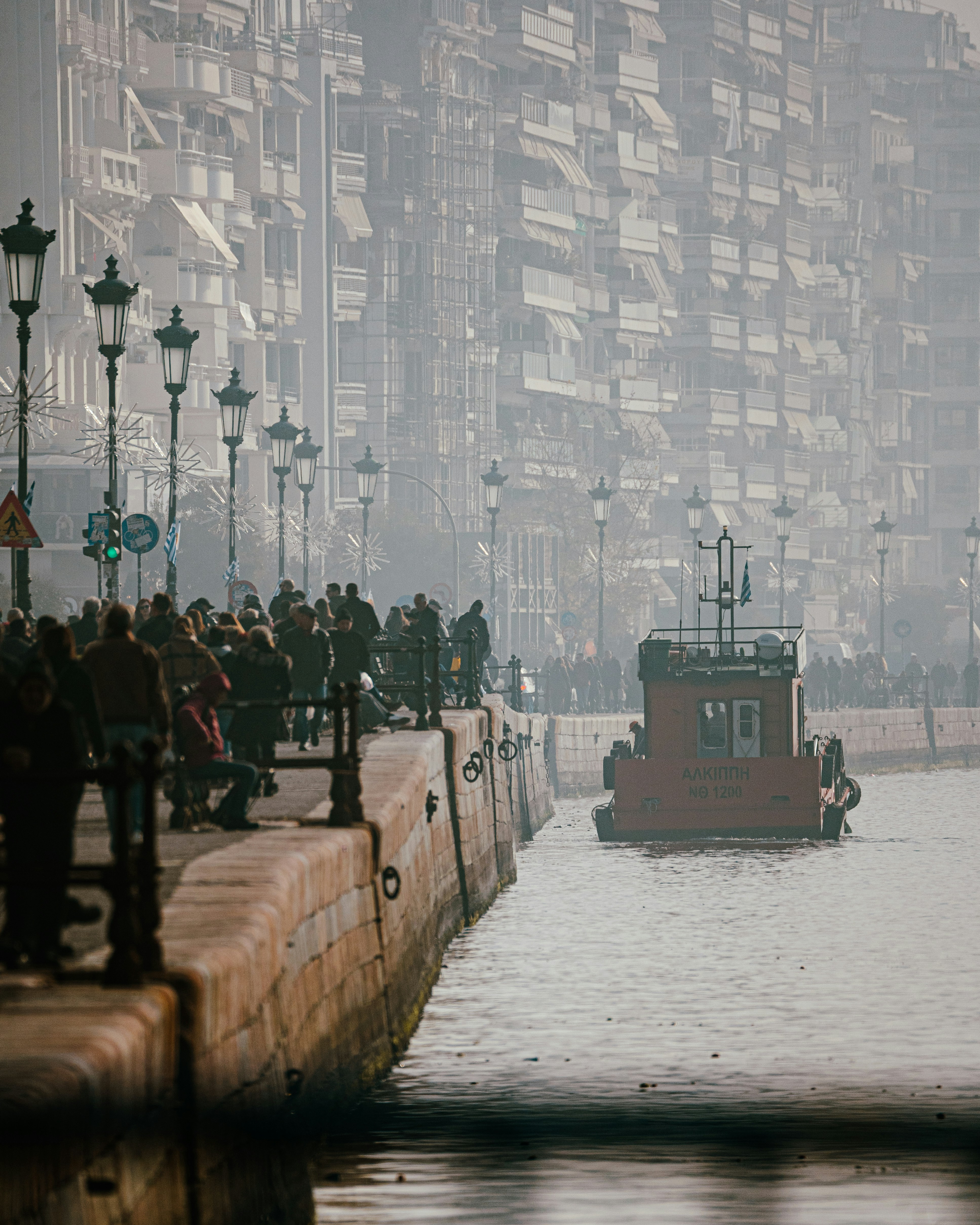Un pequeño barco rojo navega junto a un paseo marítimo.