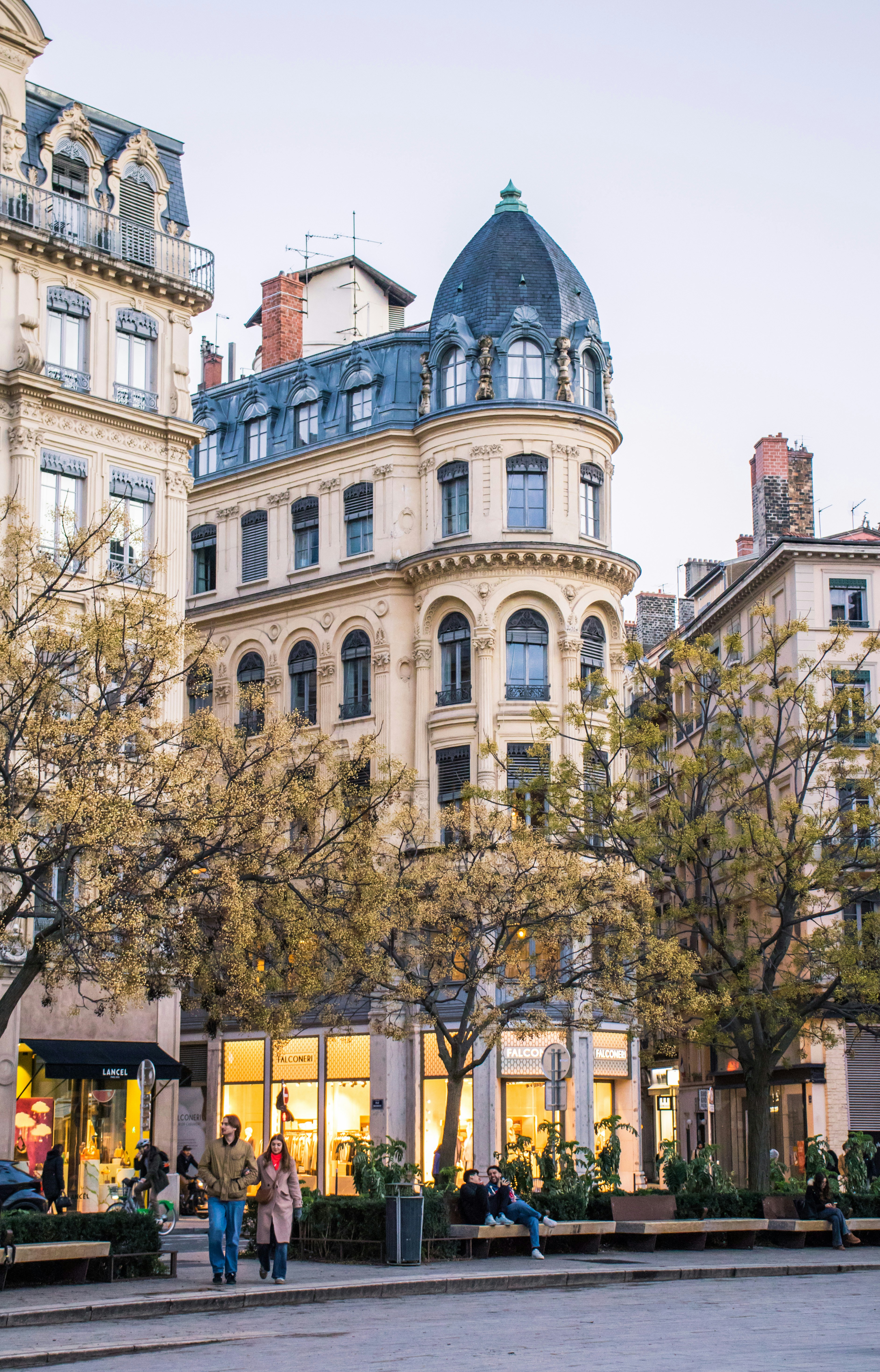 Ornate buildings line a street with trees and people.