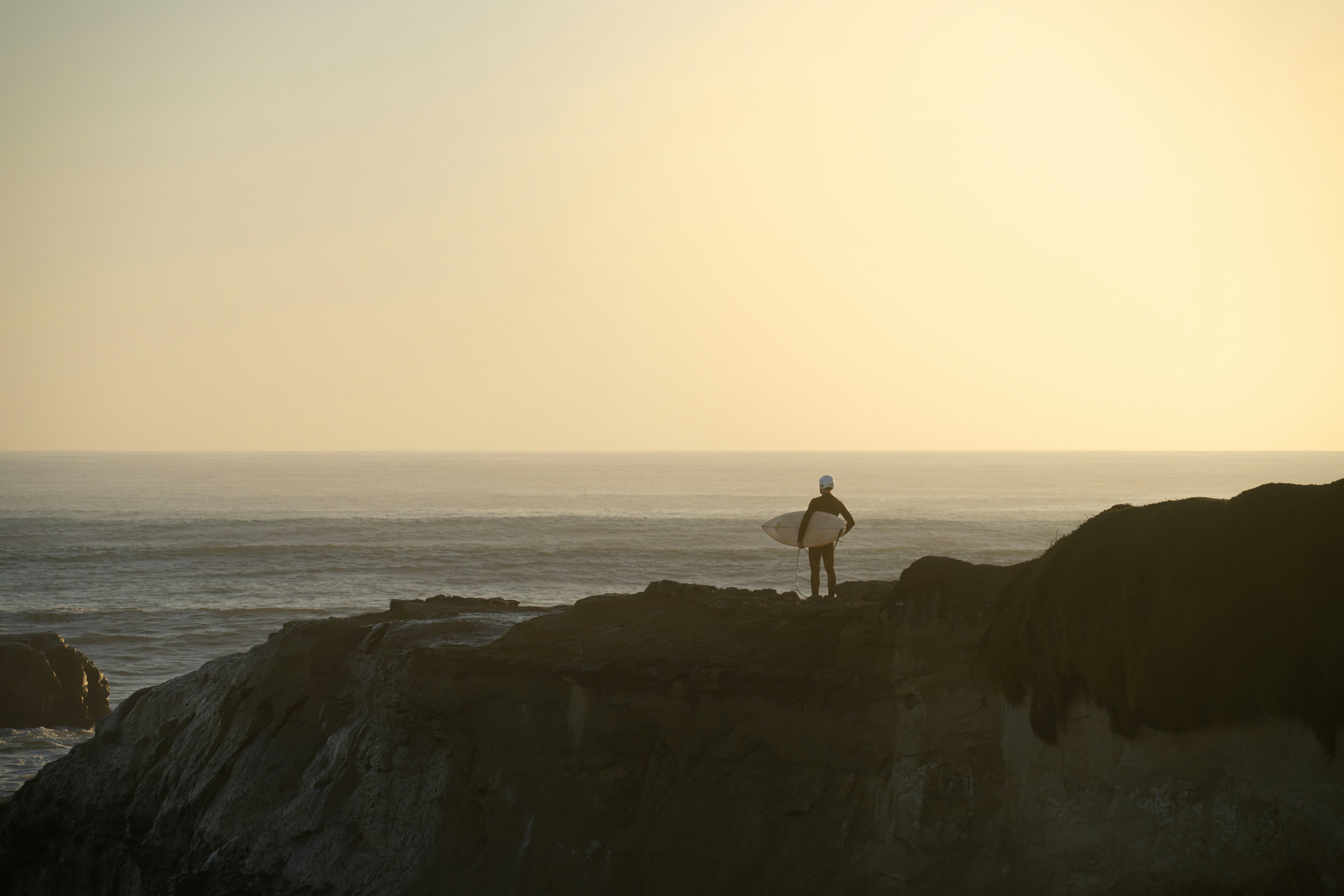 Surfer with surfboard on cliff overlooking ocean at sunset