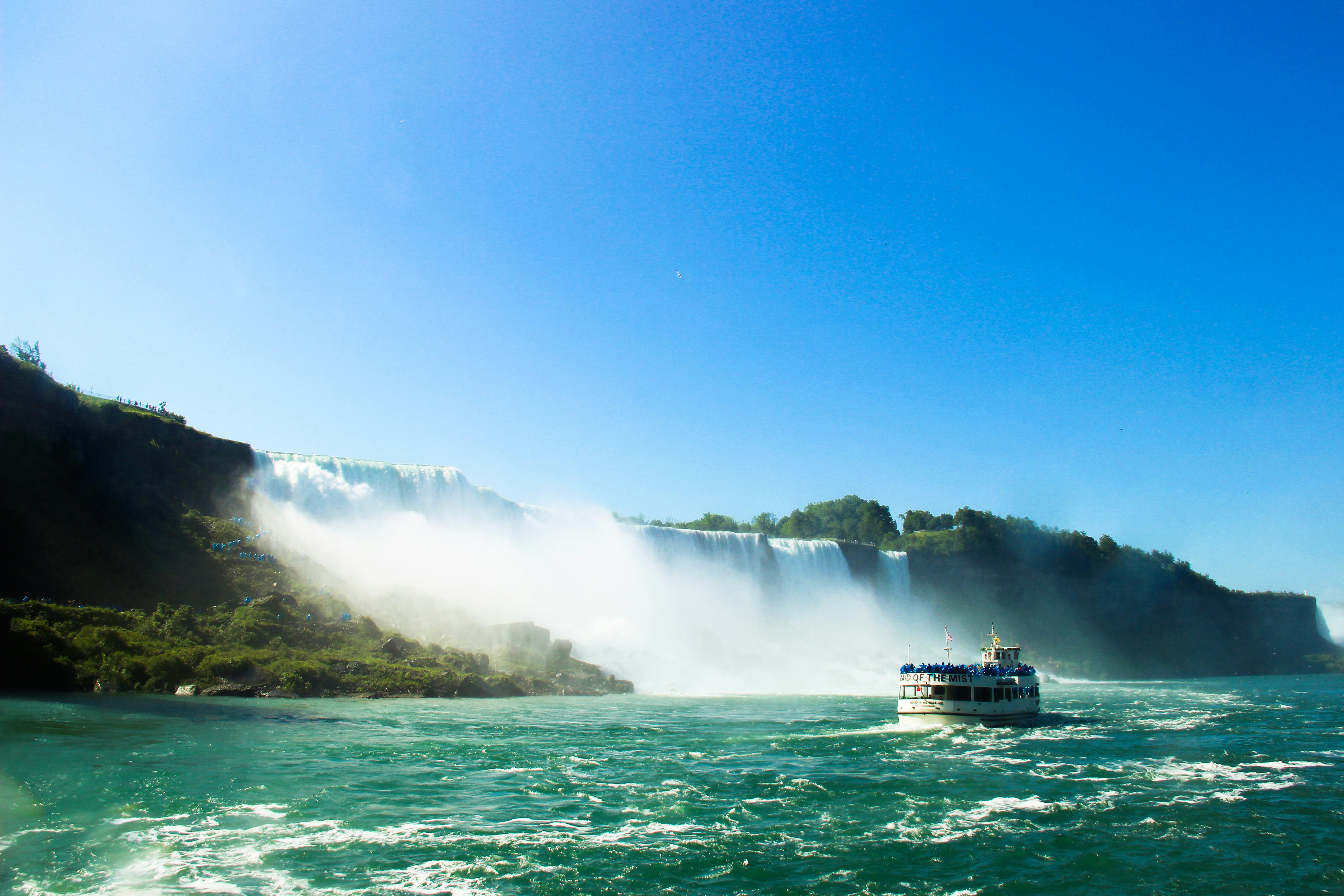 A boat approaches a misty waterfall on a sunny day