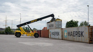 A yellow reach stacker lifts a shipping container.
