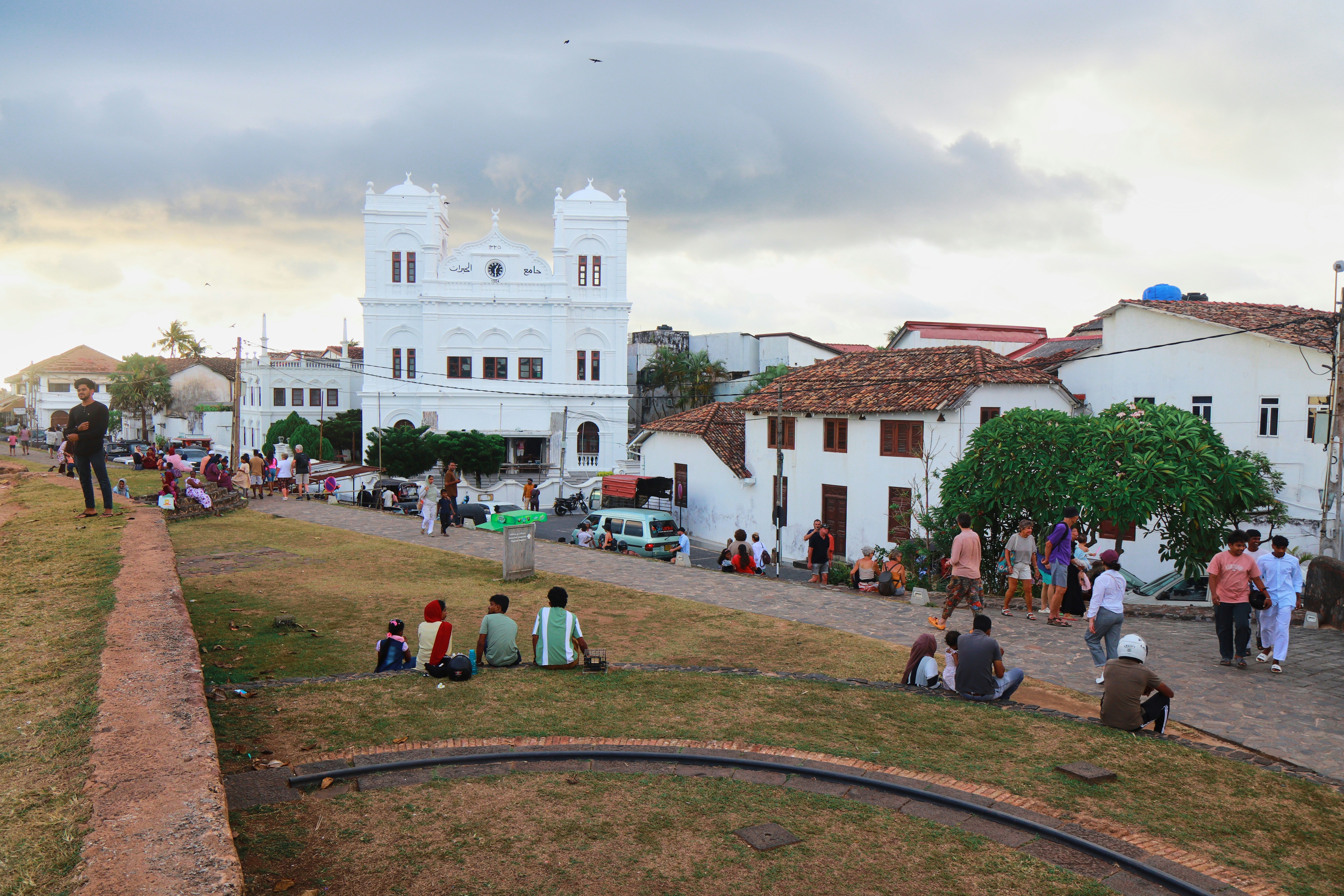 People relaxing on grassy area near white historic buildings.