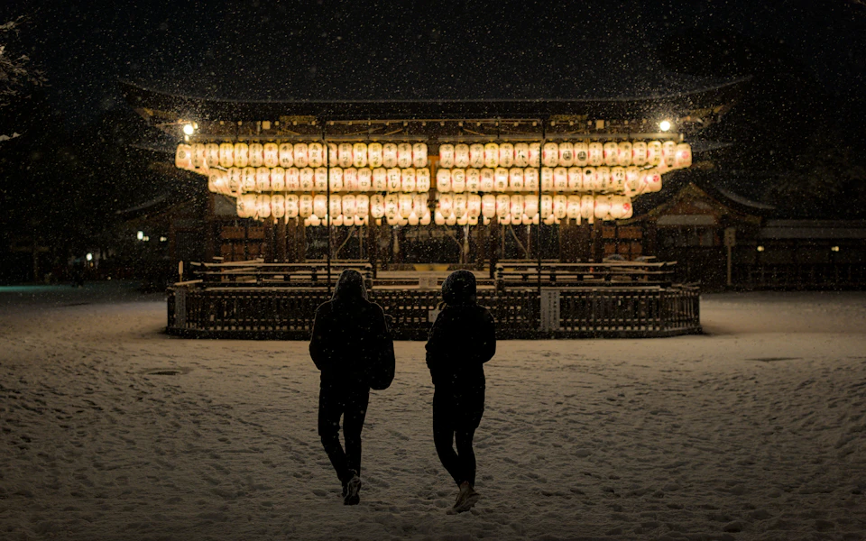 Two people walking across fresh snow towards the lantern-lit dance stage of Yasaka Shrine in Kyoto at night, with snow falling