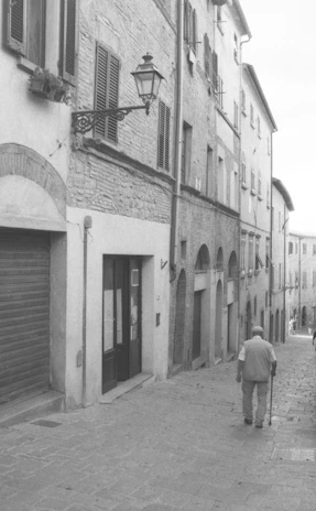Elderly man walks down a cobblestone street with buildings.