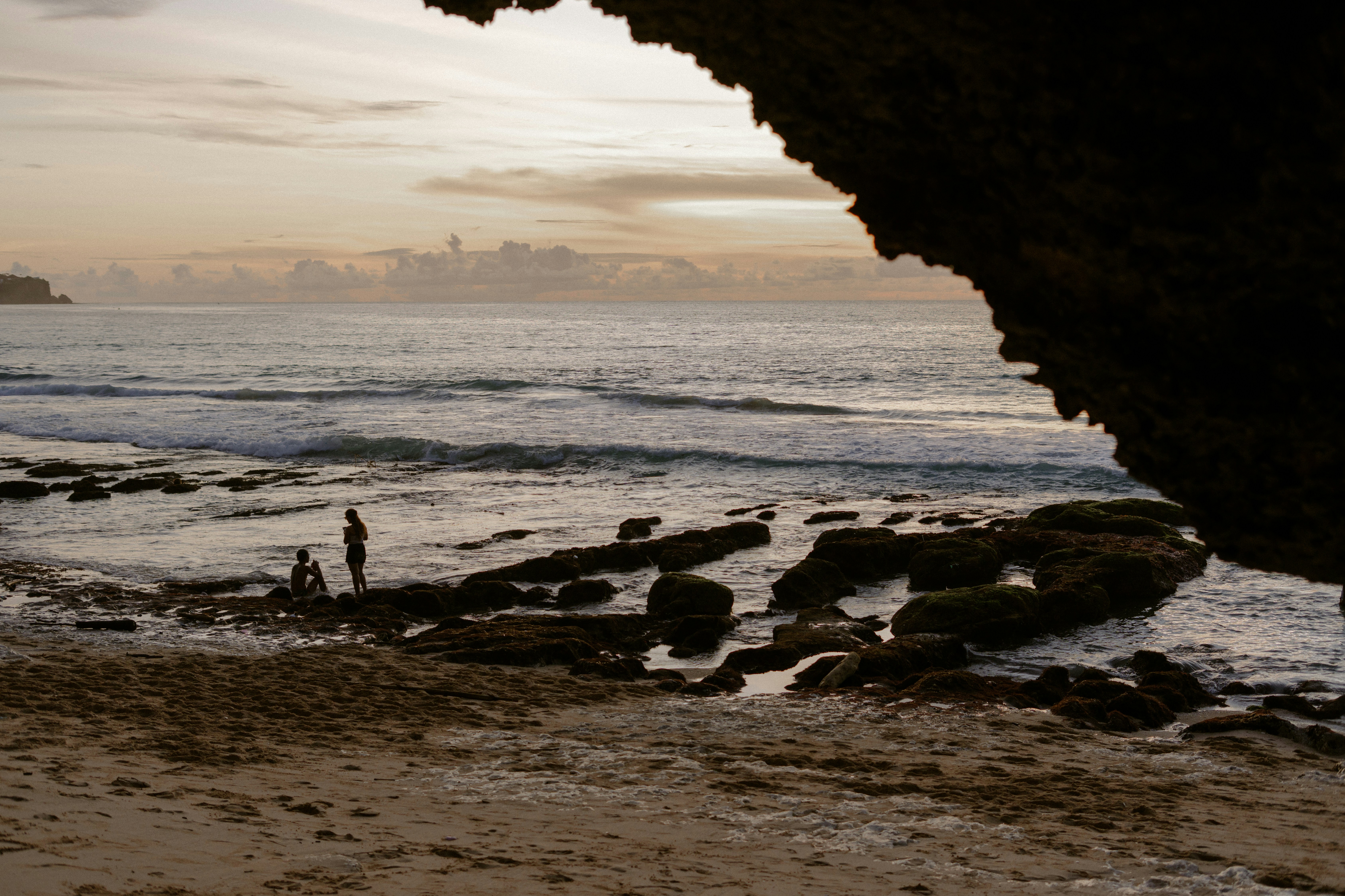 Duas pessoas estão em uma praia rochosa ao pôr do sol.