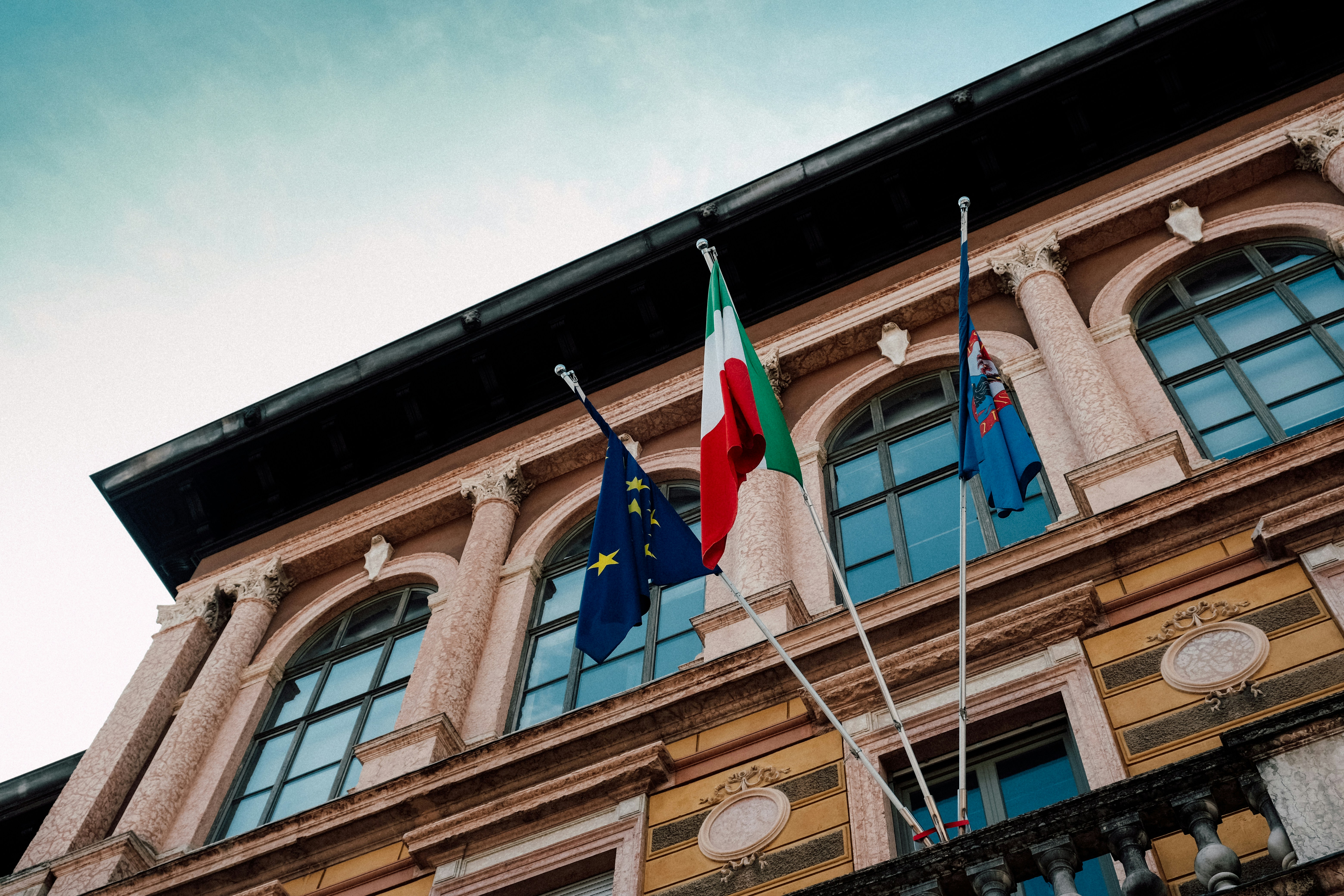 Italian and eu flags fly in front of building.