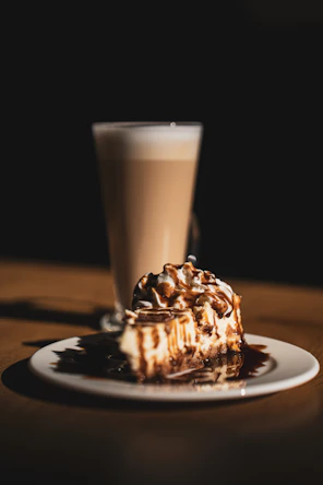 A slice of cheesecake with chocolate drizzle and latte.