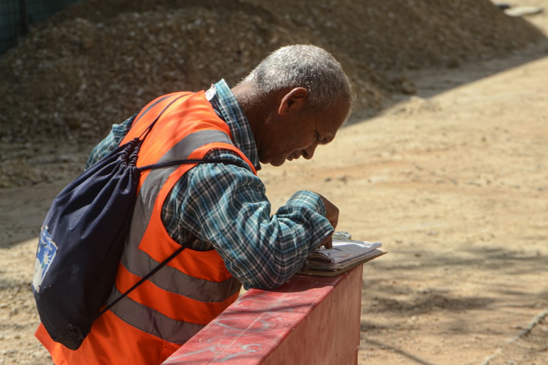 Field worker in an orange safety vest writing job notes on paper outdoors, the paperwork a phone app replaces