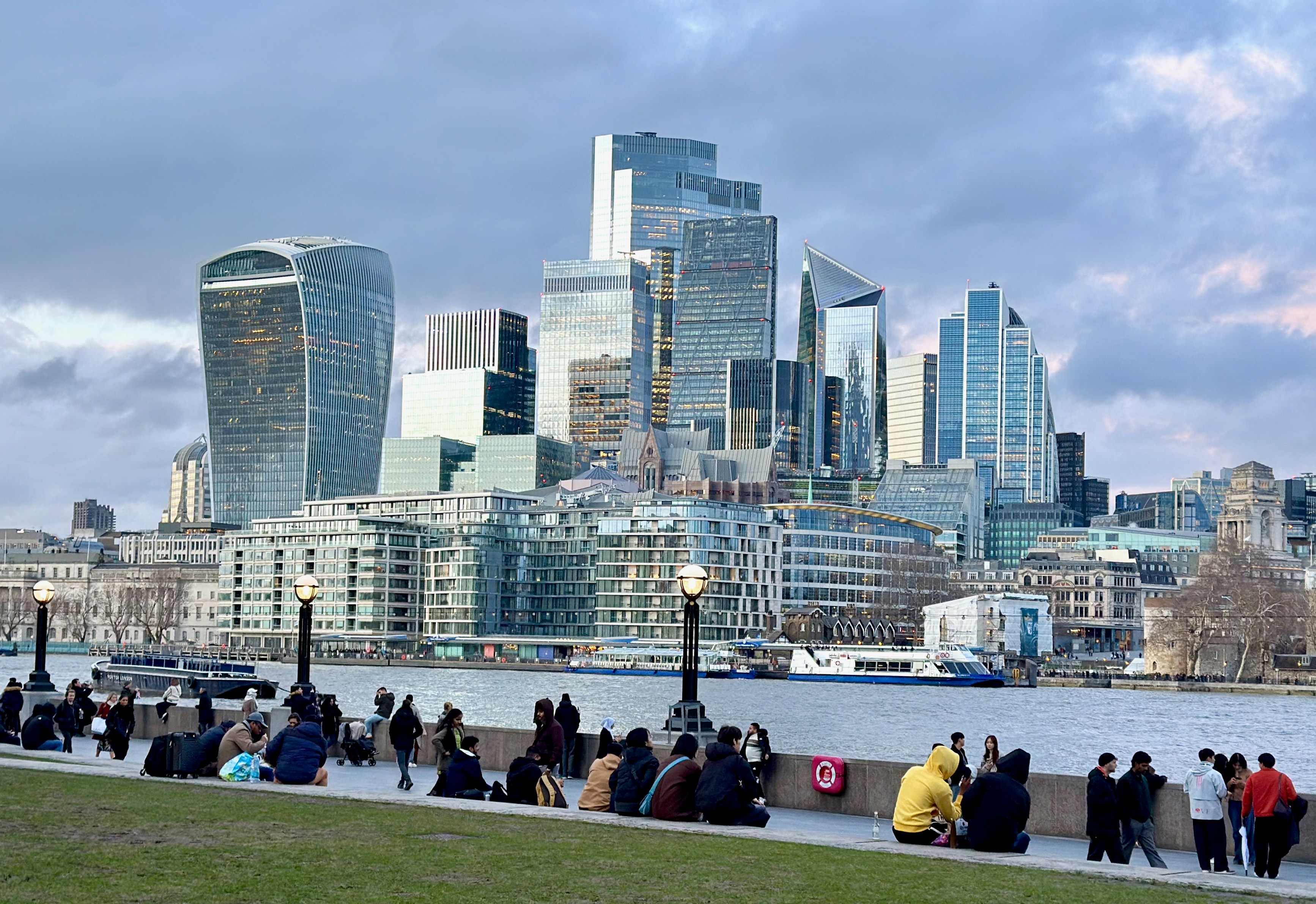 Modern skyscrapers rise above a river with people relaxing.