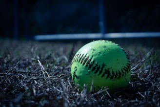A neon green softball rests on dry grass