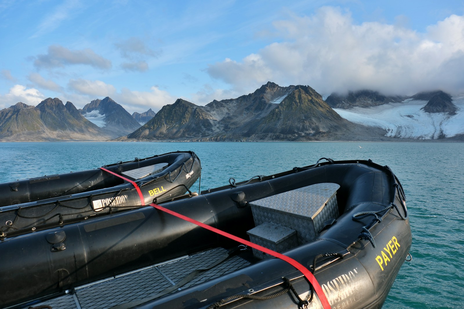 Two black zodiac boats moored together on calm water in front of a mountain range and a glacier
