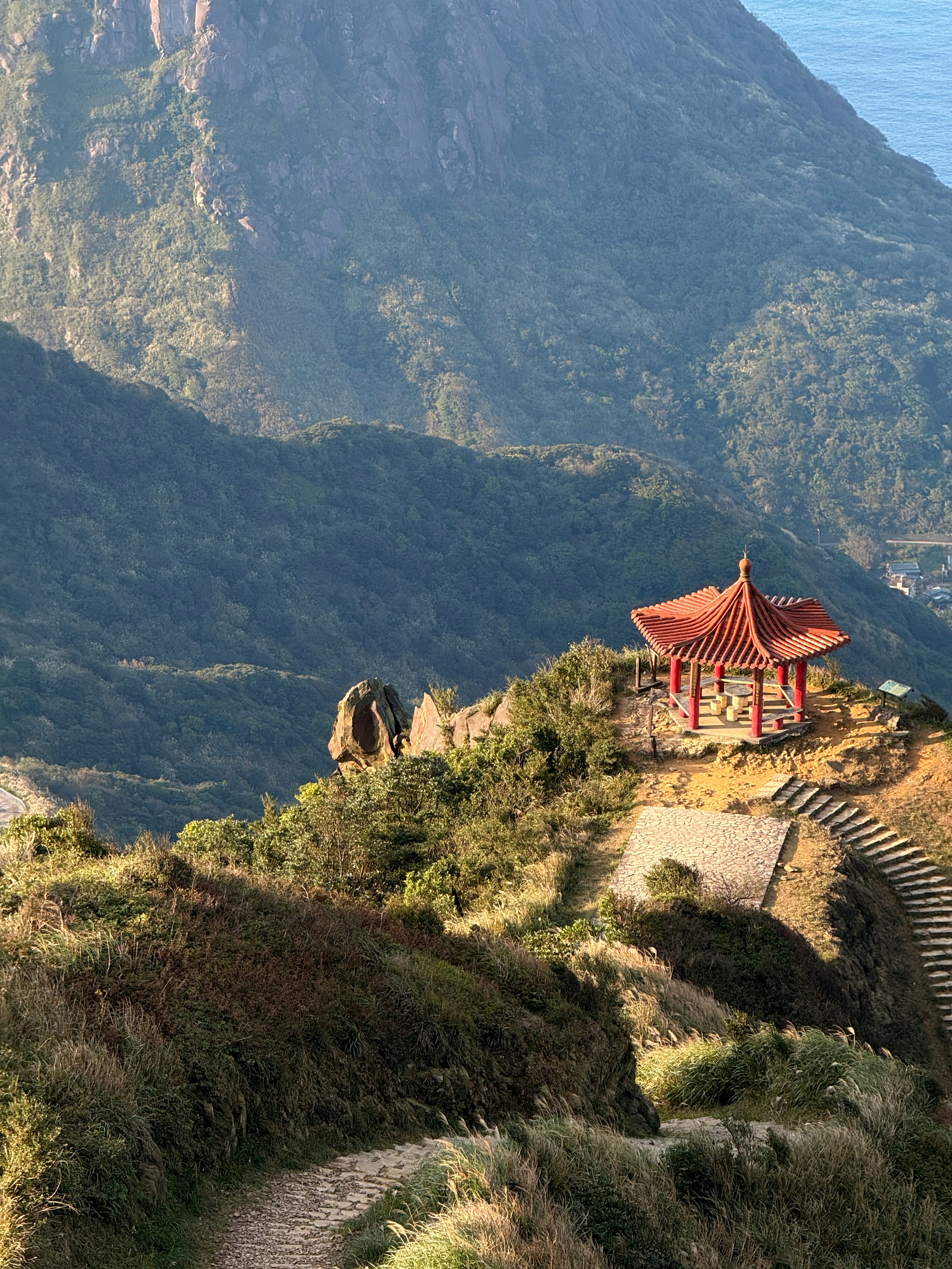 Red pagoda on a grassy mountain overlooking the ocean.