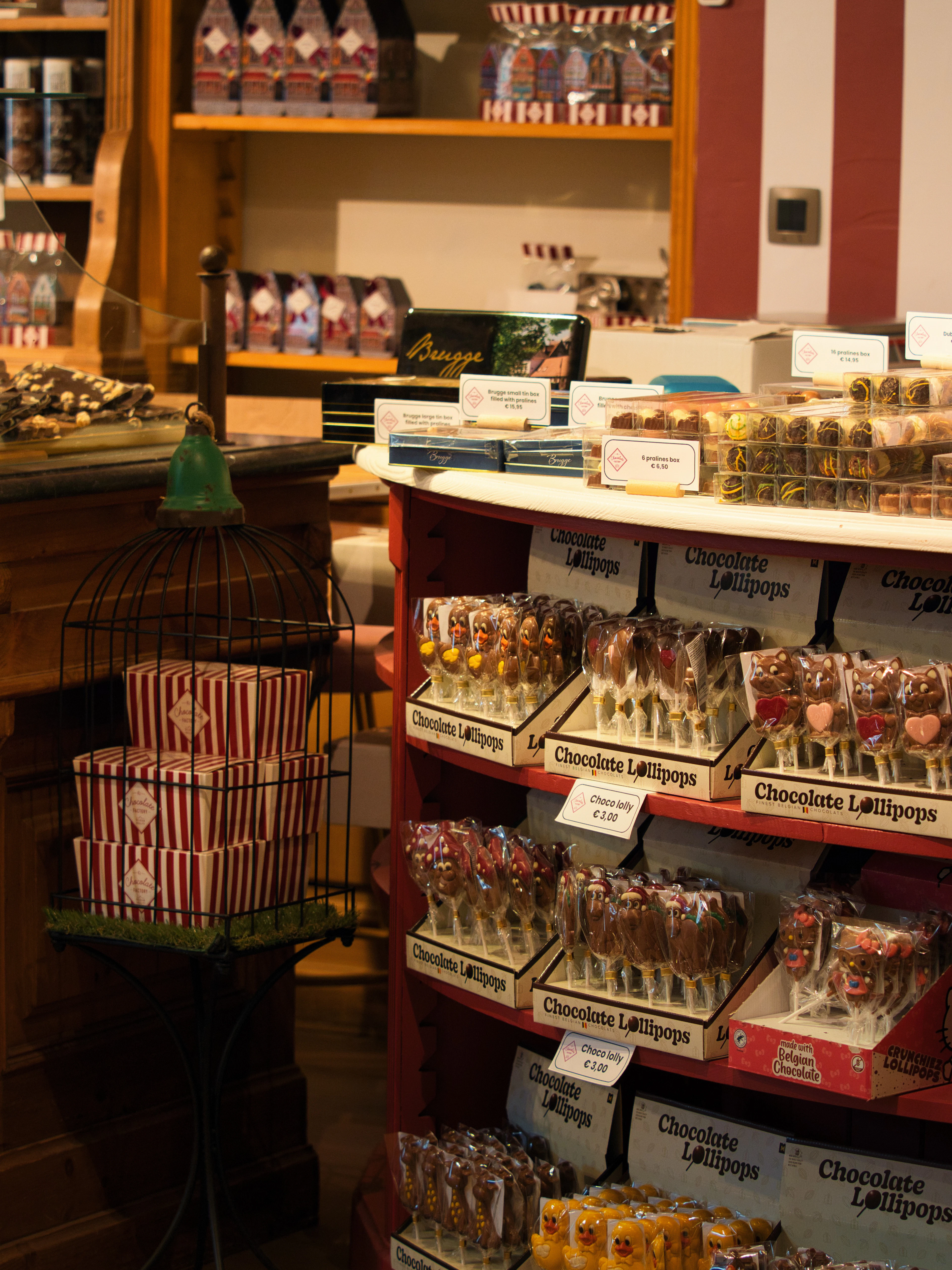Inside a chocolate shop in Bruges: tiered trays of chocolate lollipops with printed price cards, boxed pralines and striped candy tins on the wooden shelving behind, and a small script sign reading 'Bruges' on the counter.