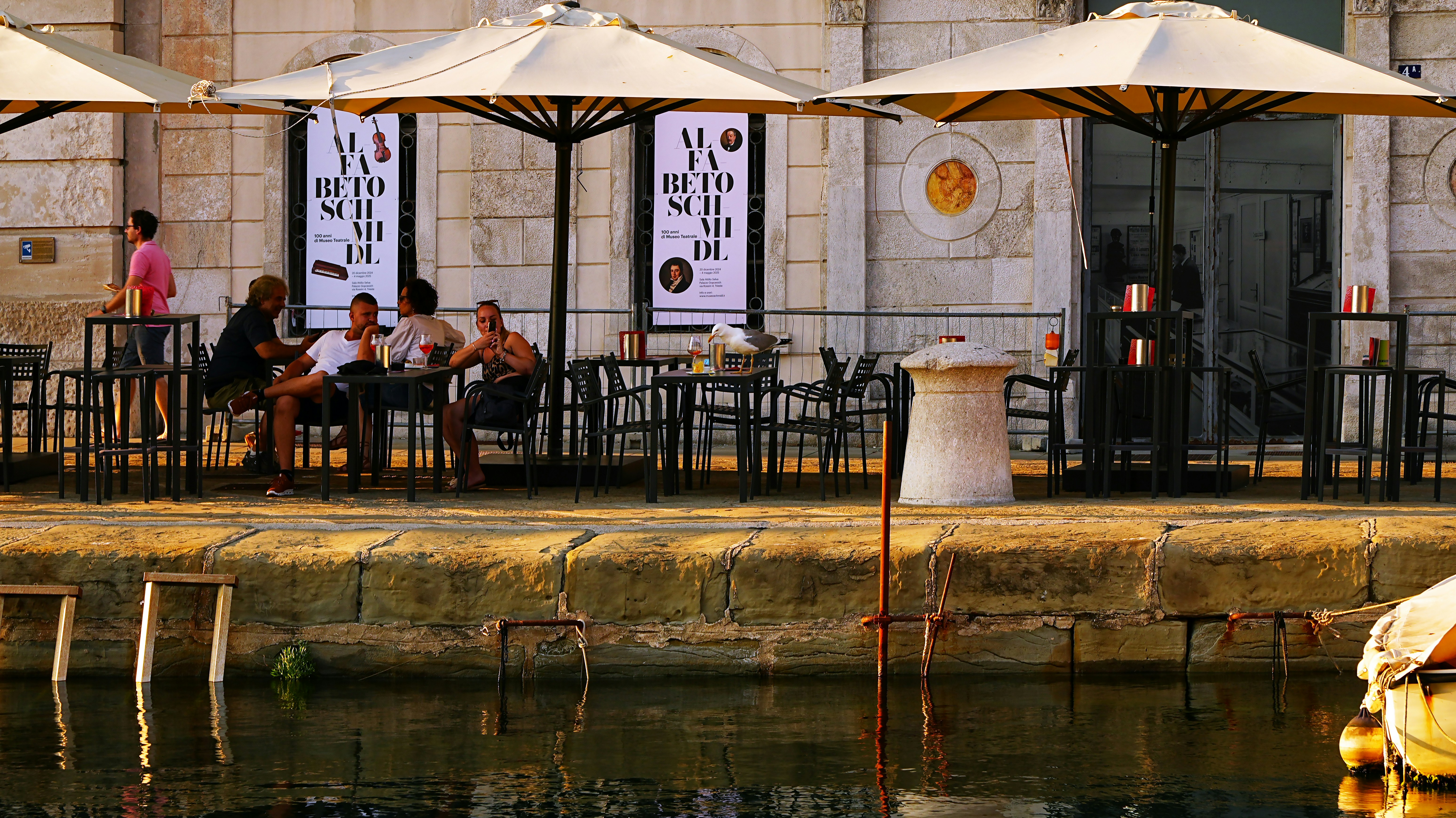 Die Leute sitzen an Tischen unter Regenschirmen vor einem Gebäude.