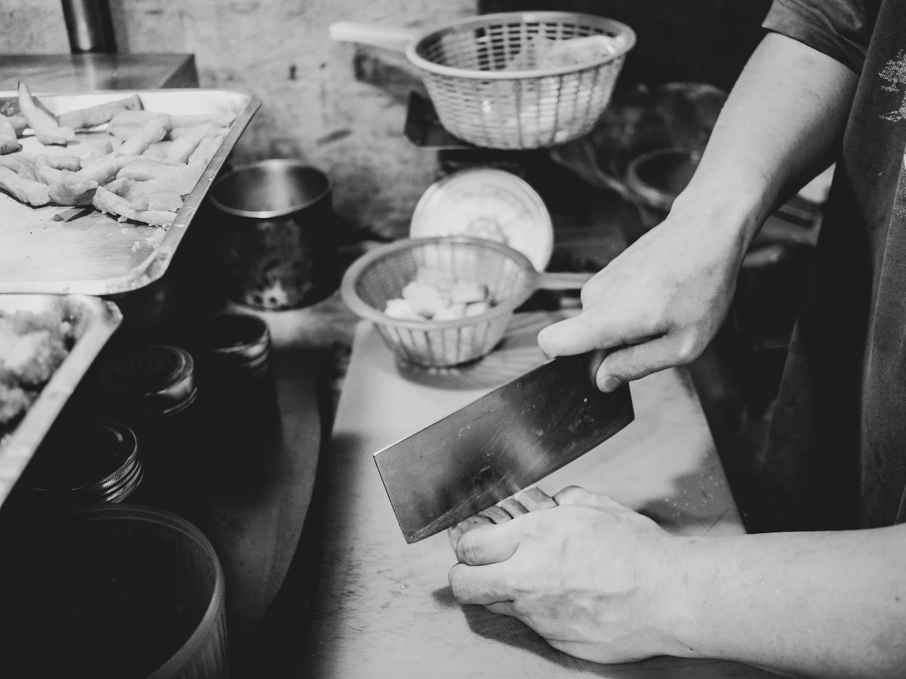 Black-and-white photo of hands slicing with a Chinese cleaver, prep baskets behind