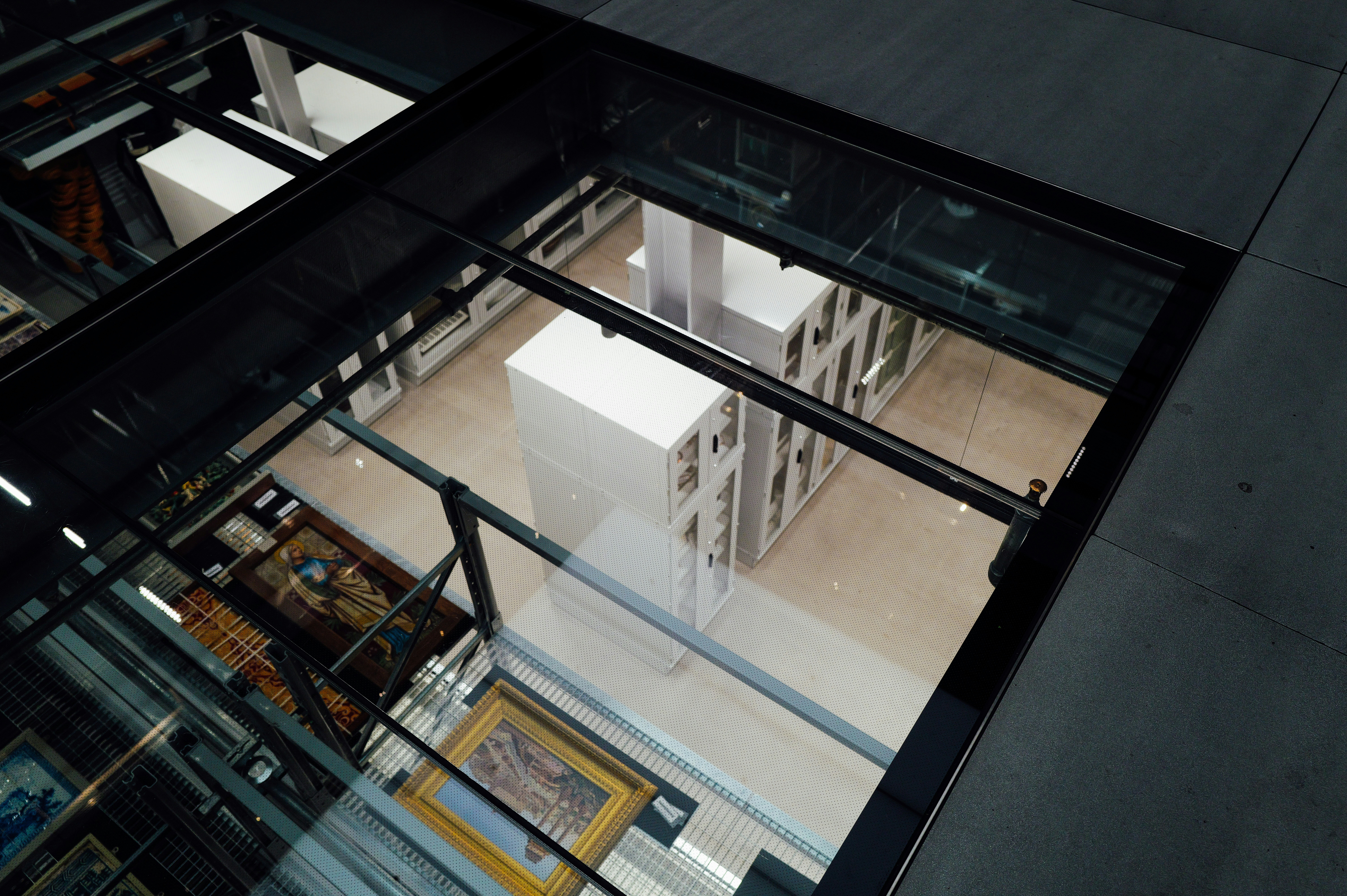 An overhead view through a dark-framed glass floor reveals a modern museum storage area below, with white cases, art, and light floors.