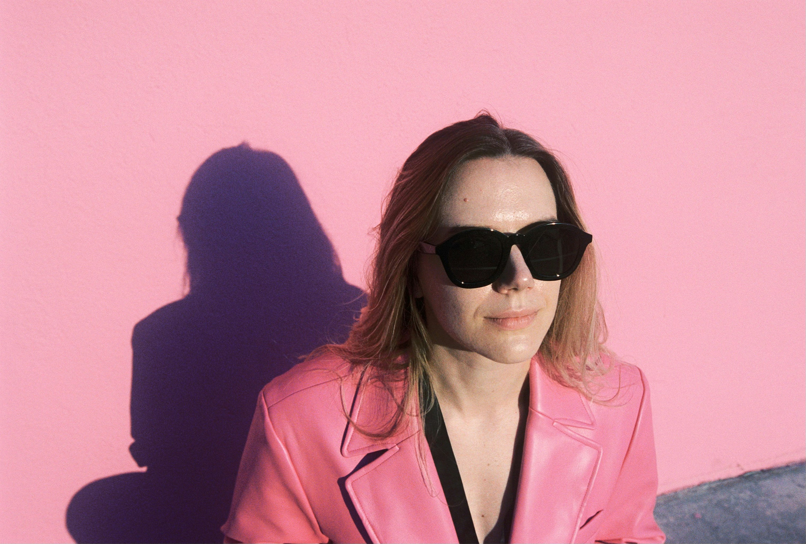 Mulher de blazer rosa e óculos escuros contra a parede rosa.