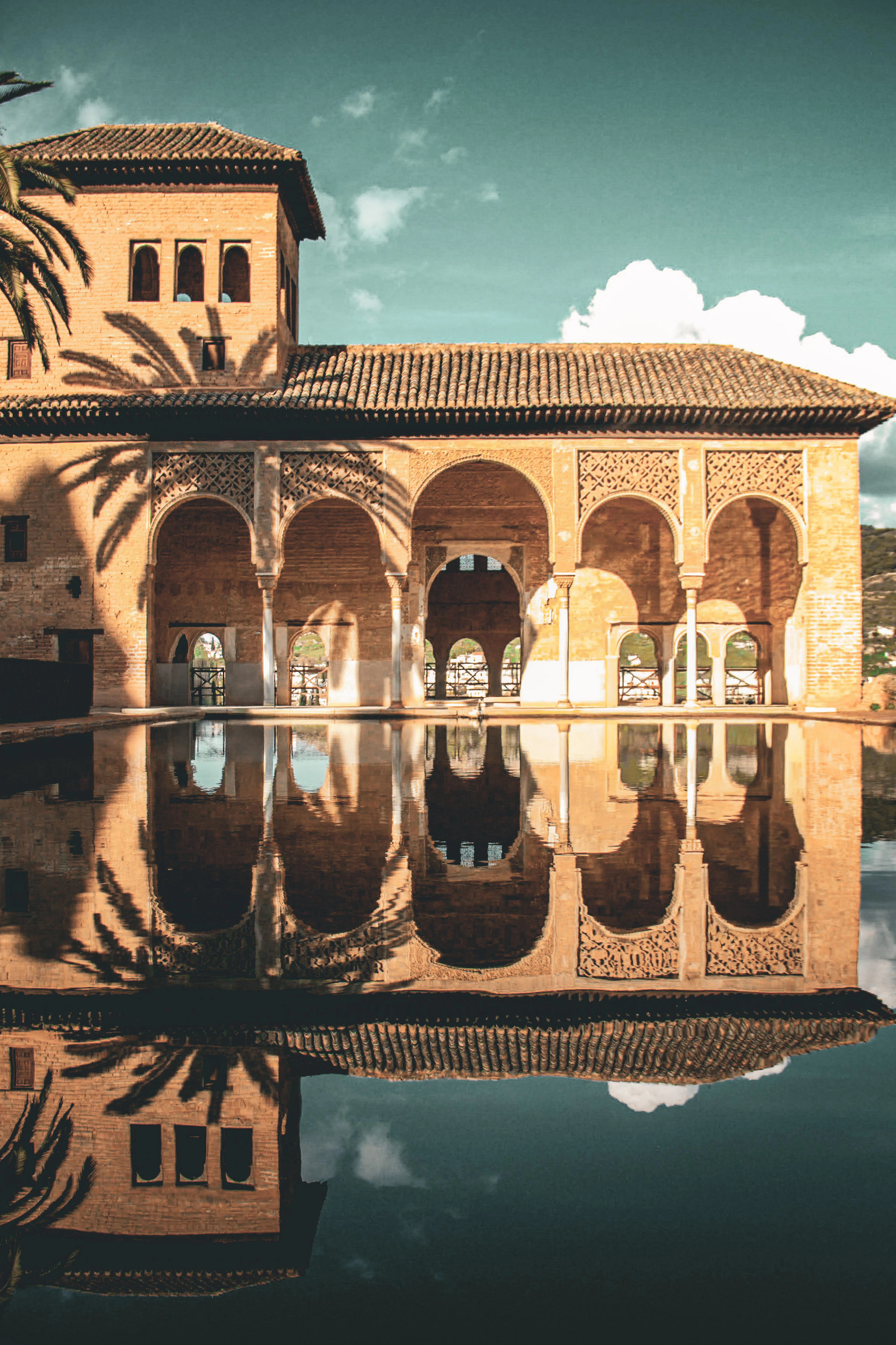 The arcaded Partal Palace at the Alhambra in Granada mirrored in its still reflecting pool, warm stone and carved plasterwork under a palm frond's shadow