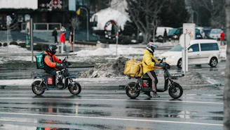 Two delivery drivers on scooters on a wet street