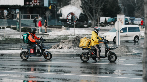 Two delivery drivers on scooters on a wet street