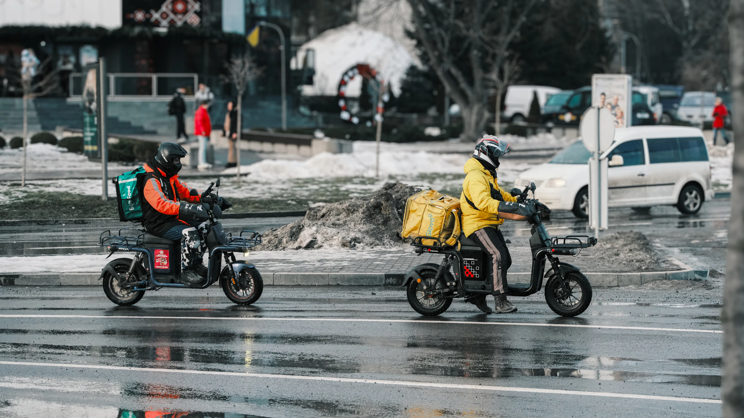 Deux livreurs 2-roues avec sacs thermiques jaune et vert en circulation urbaine sous la pluie