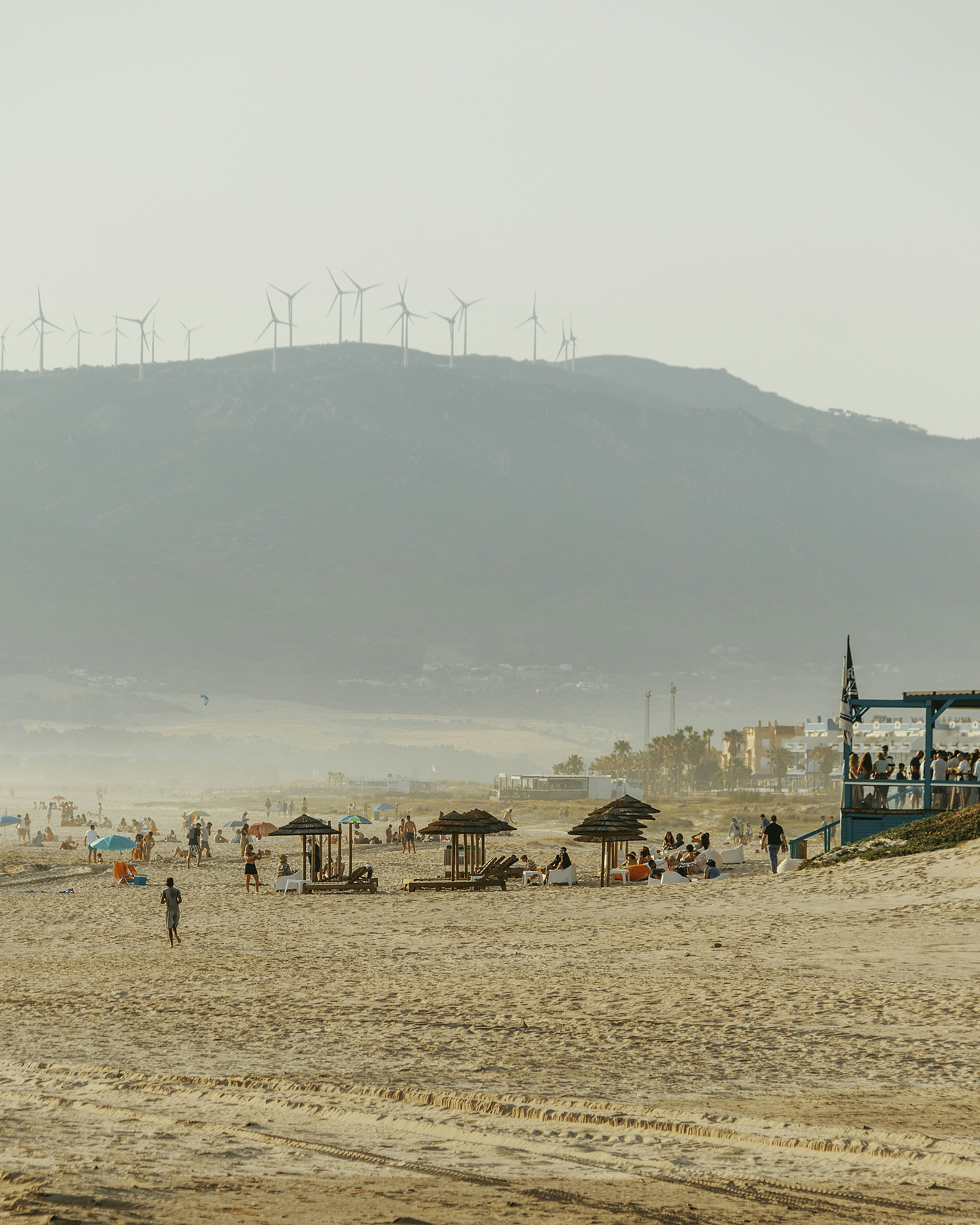 Pessoas relaxando em uma praia de areia com moinhos de vento na colina.