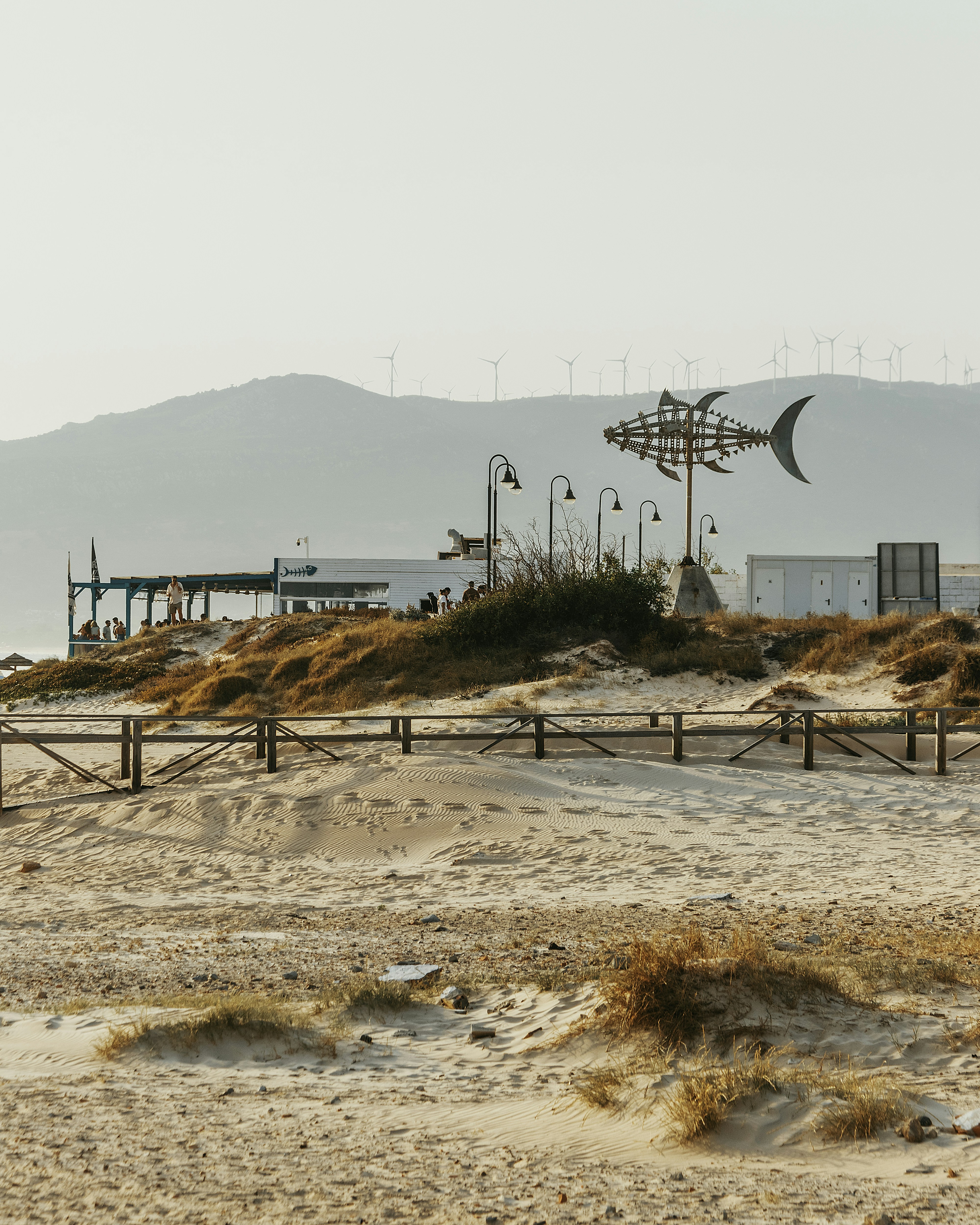 Escultura de peixe metálico em uma praia de areia com prédios.