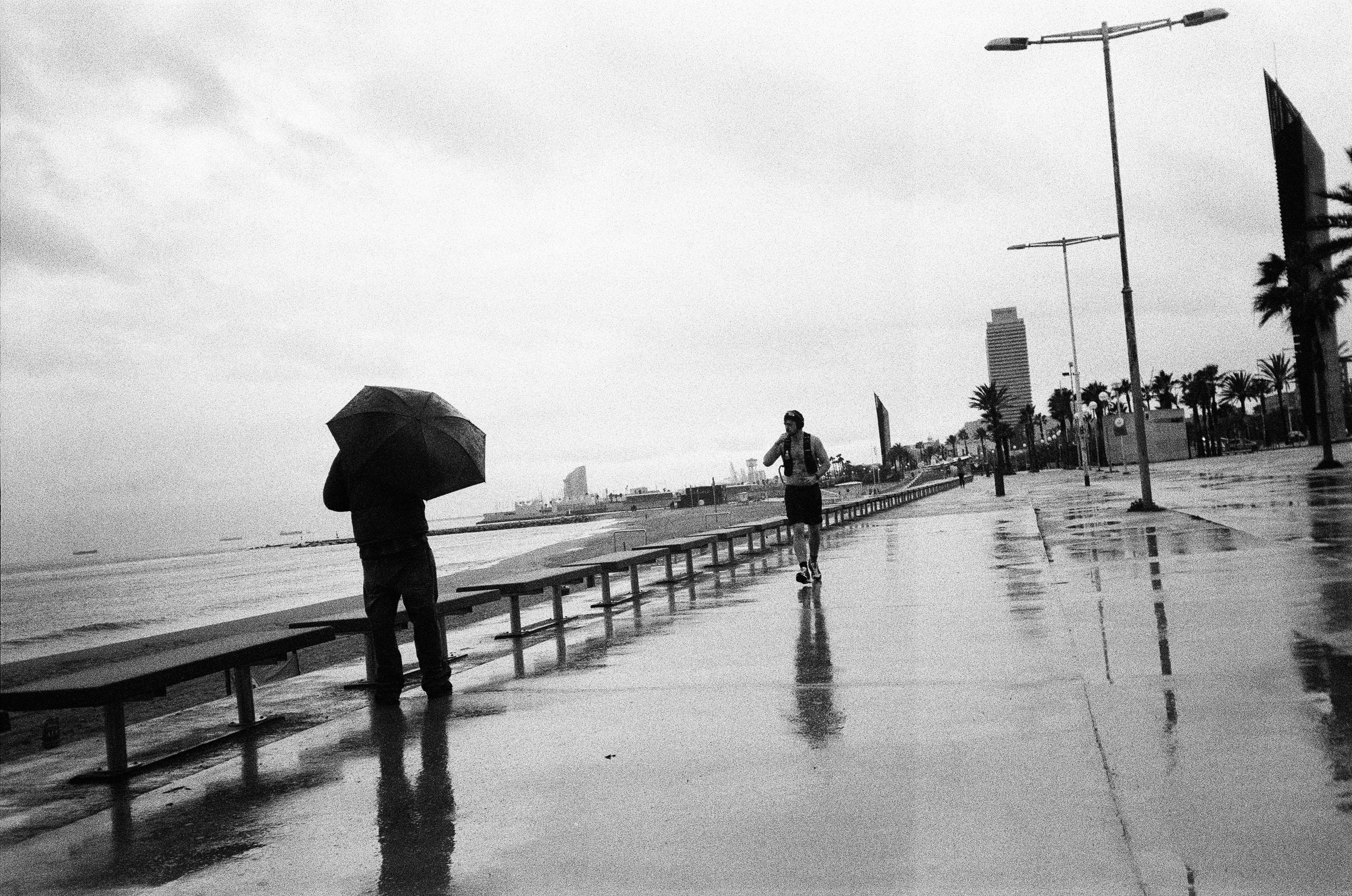 Man with umbrella watches runner on wet promenade