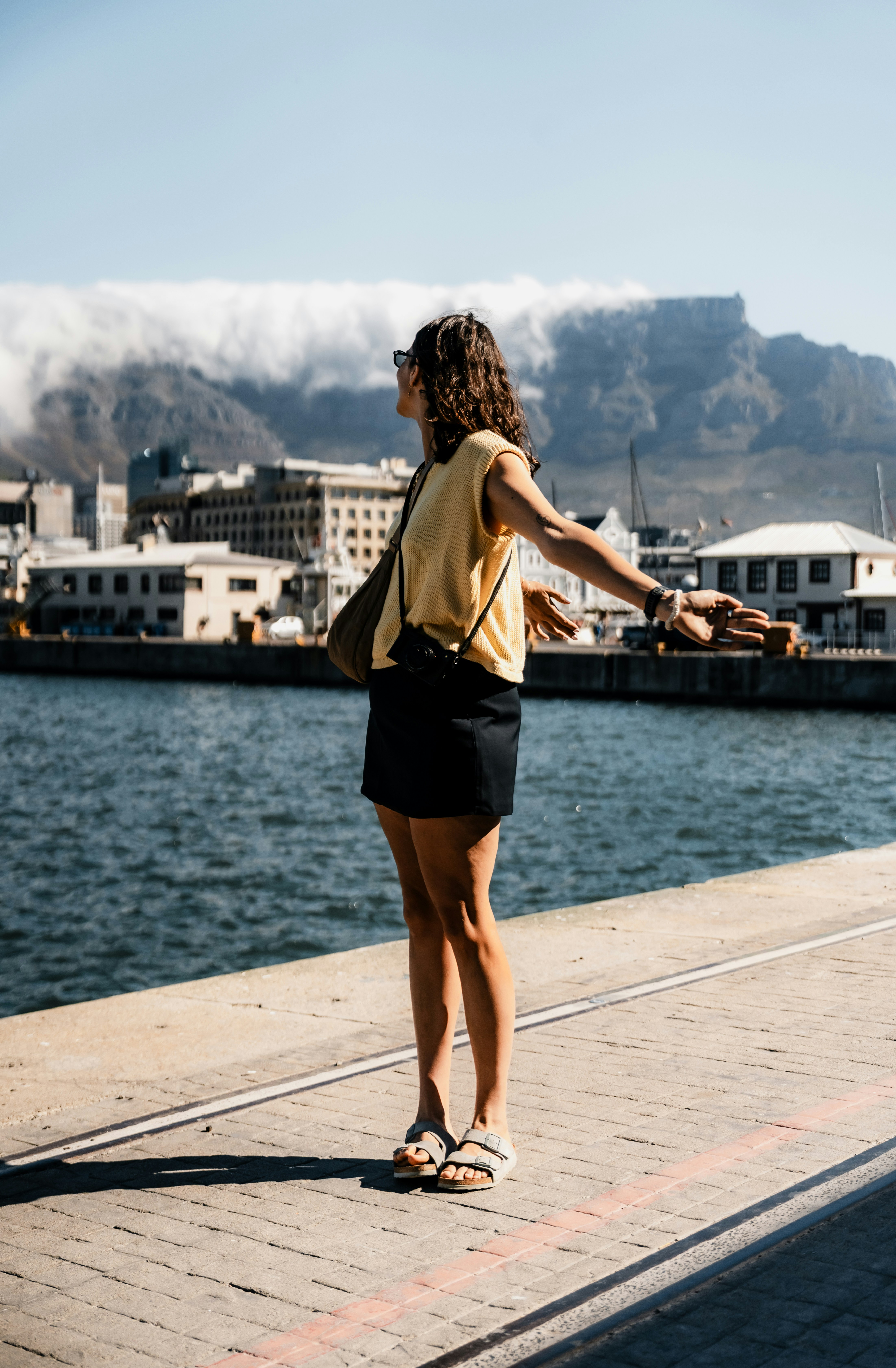 Mujer con los brazos extendidos junto al agua y montañas detrás.