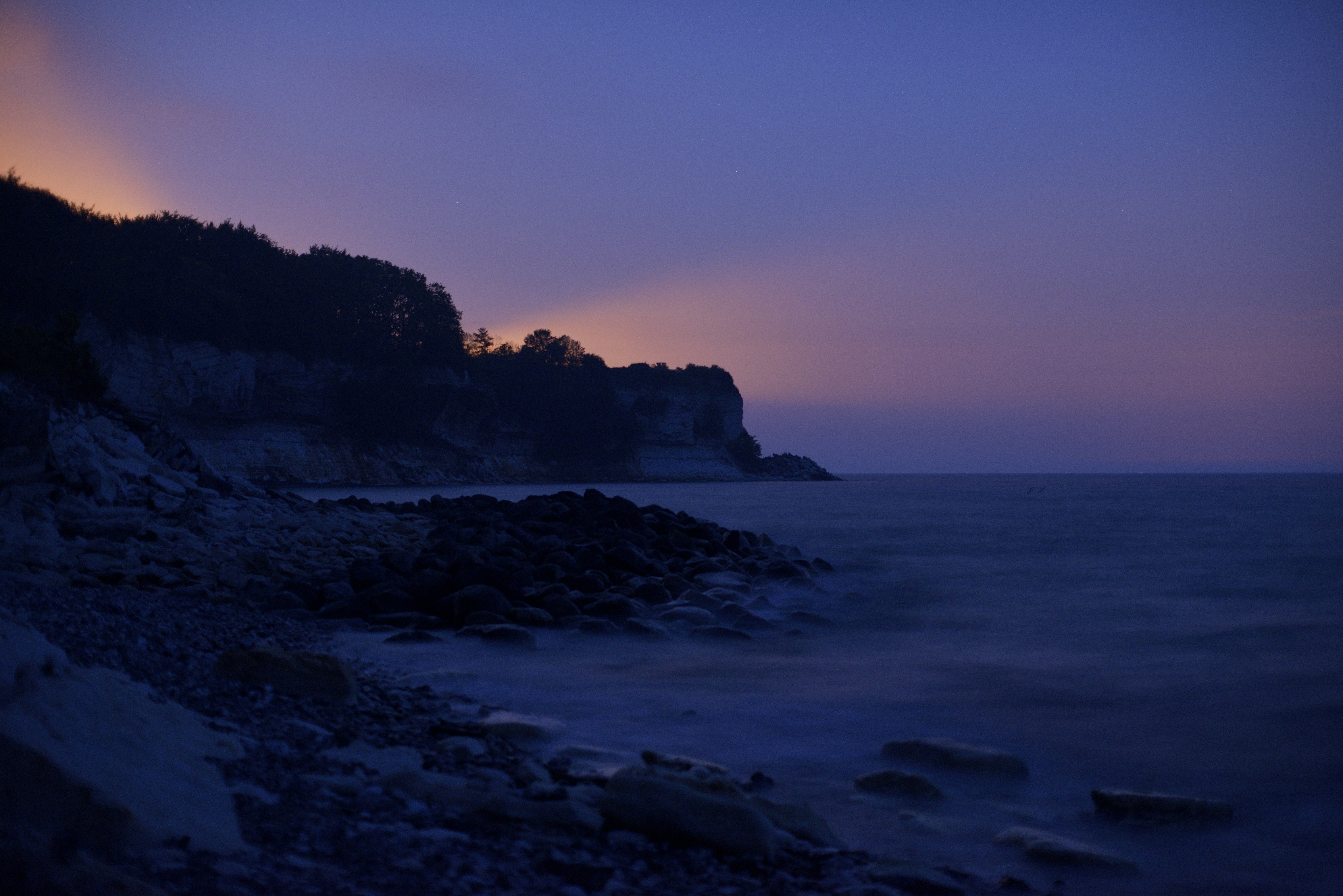 Dark ocean coastline at twilight with distant light.