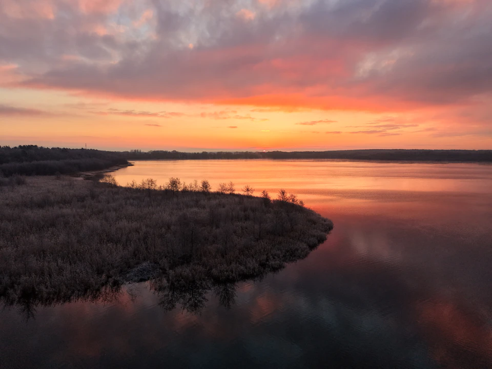 The image depicts a serene lake at sunset, with the sky painted in hues of orange and pink. The sun is setting behind the horizon, casting a warm glow over the scene. The calm water reflects the vibrant colors of the sky, creating a beautiful mirror-like effect. On one side of the lake, there are trees and bushes that add to the natural beauty of the landscape.