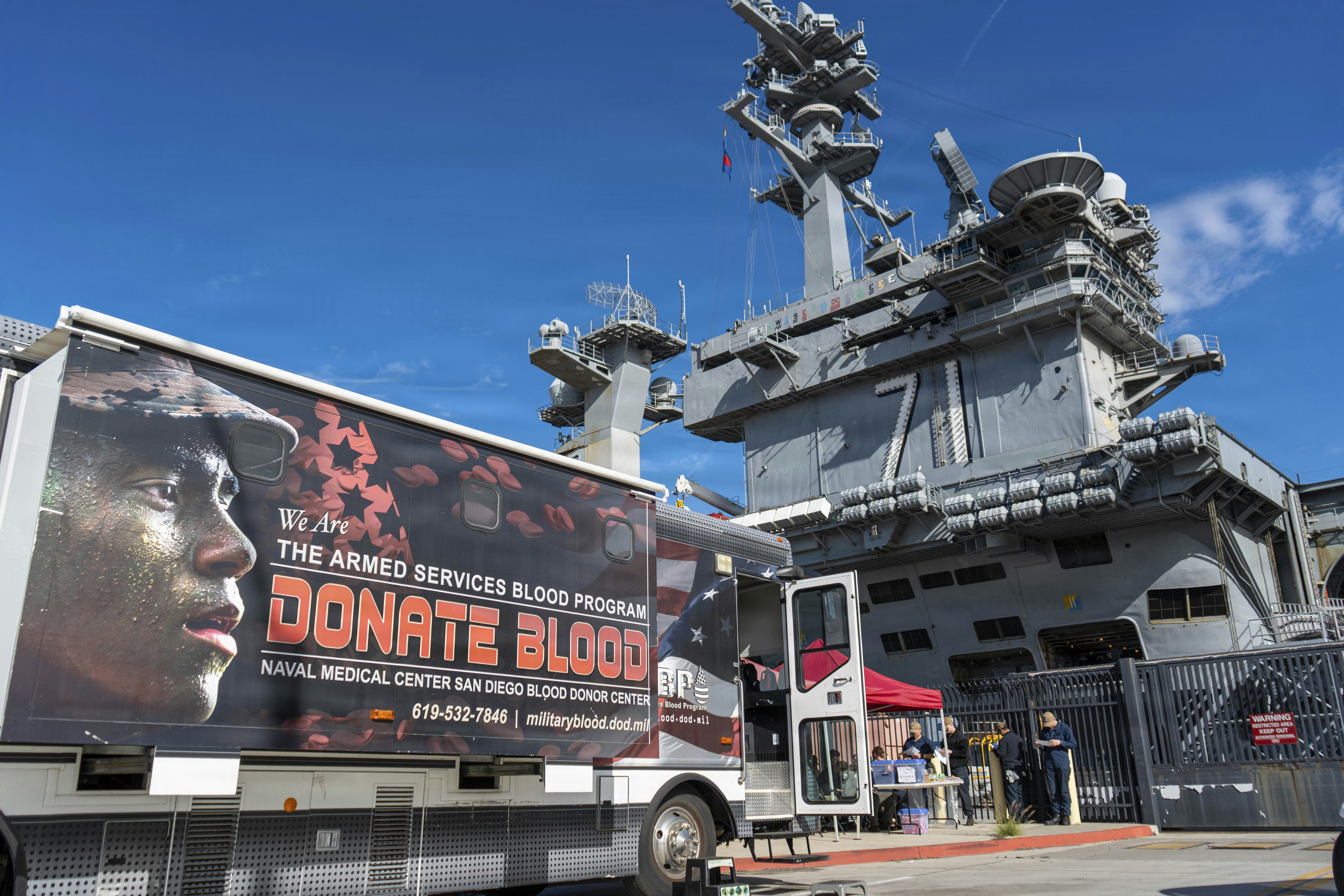 Camión de donación de sangre aparcado cerca de un gran portaaviones.