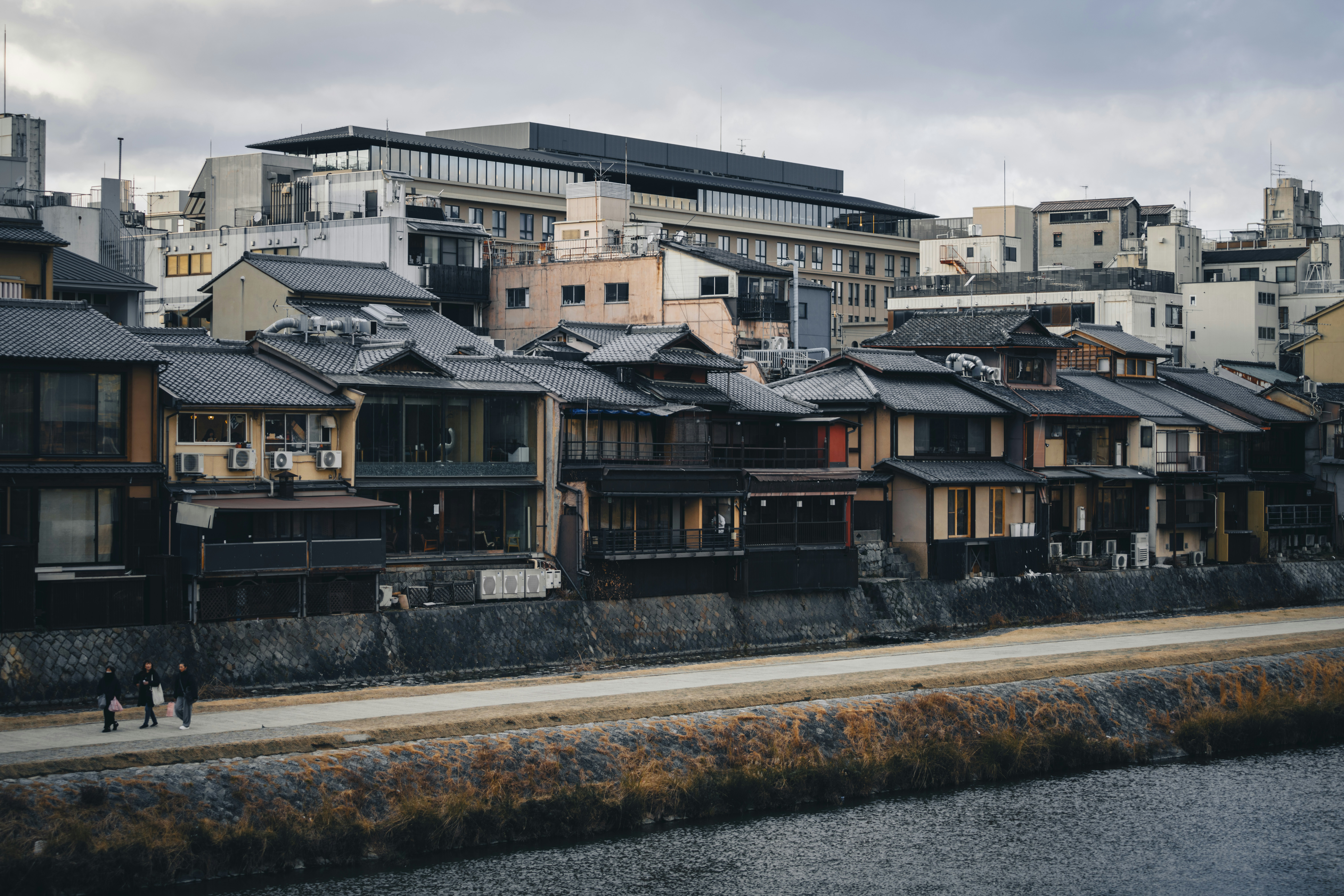 Traditional japanese buildings line a riverbank under cloudy skies.