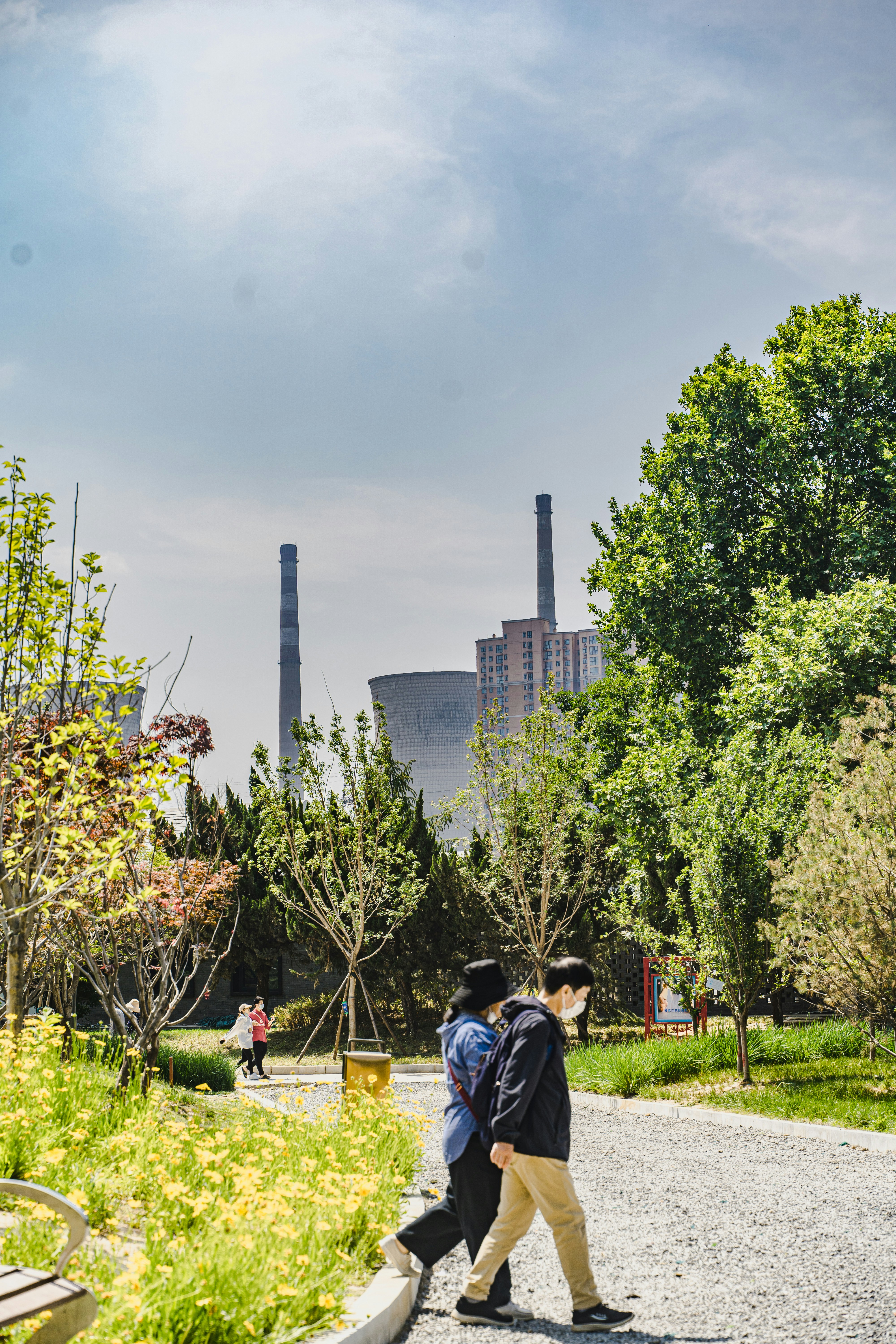 Die Leute gehen an Bäumen vorbei, auf denen im Hintergrund Industrietürme stehen.