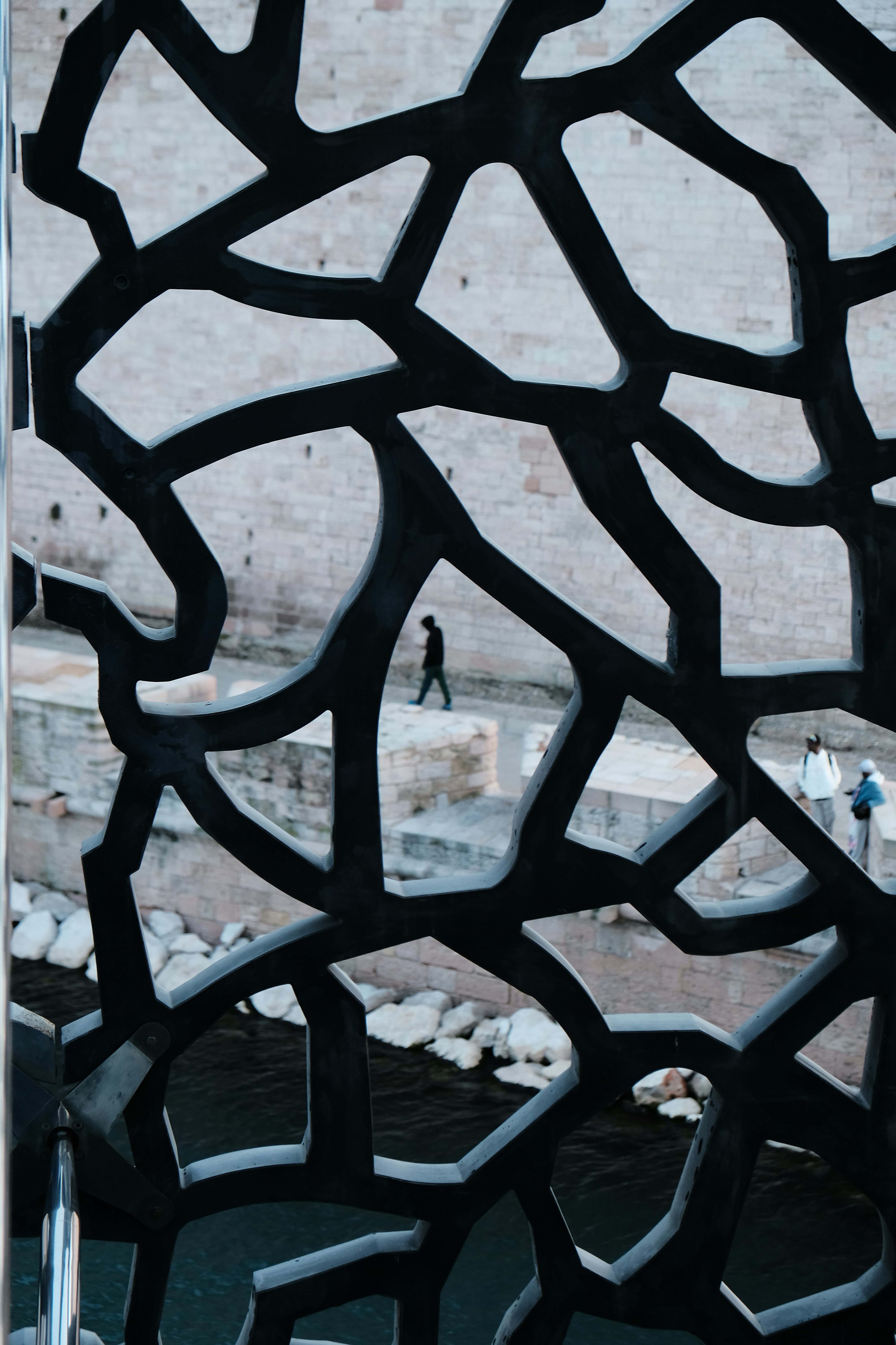 Pessoas passando por uma estrutura metálica com padrão perto da água.