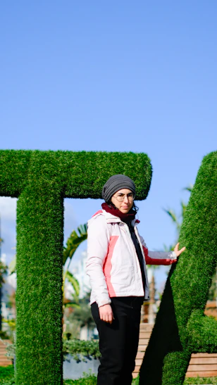 Woman in hat stands by large green hedge letters