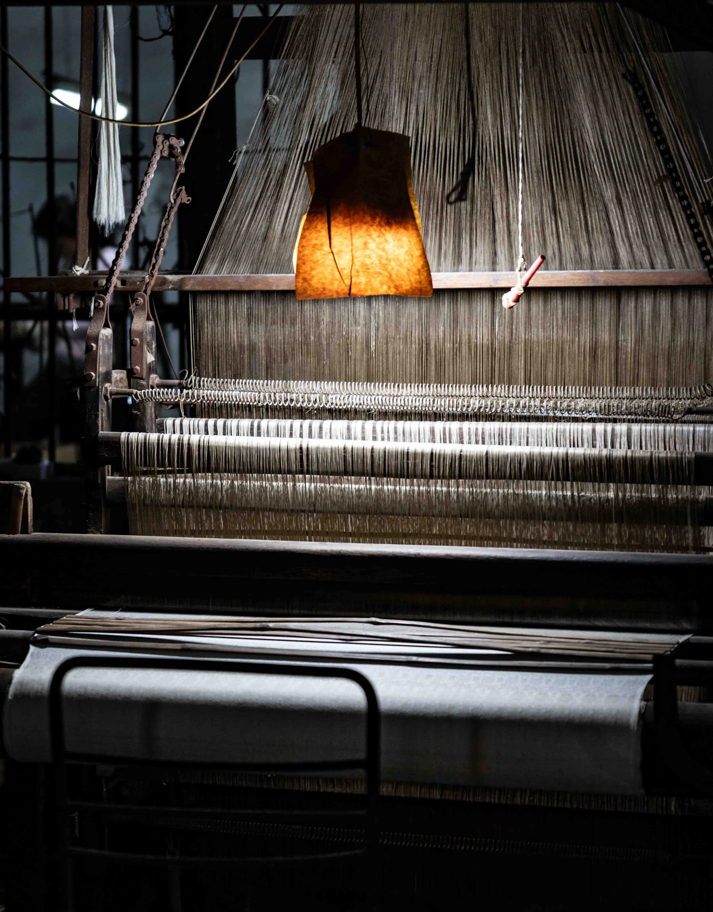 Vertical loom warp lit by a single hanging lamp inside a darkened weaving shed
