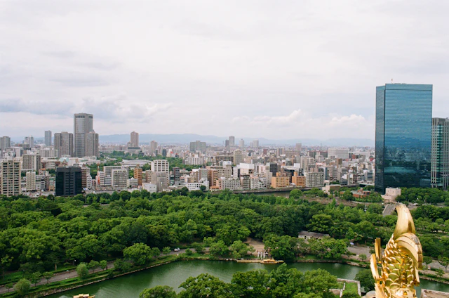 A view from Osaka Castle's observation deck over the castle park's greenery and pond toward the Osaka skyline, with a golden shachihoko roof ornament in the foreground.
