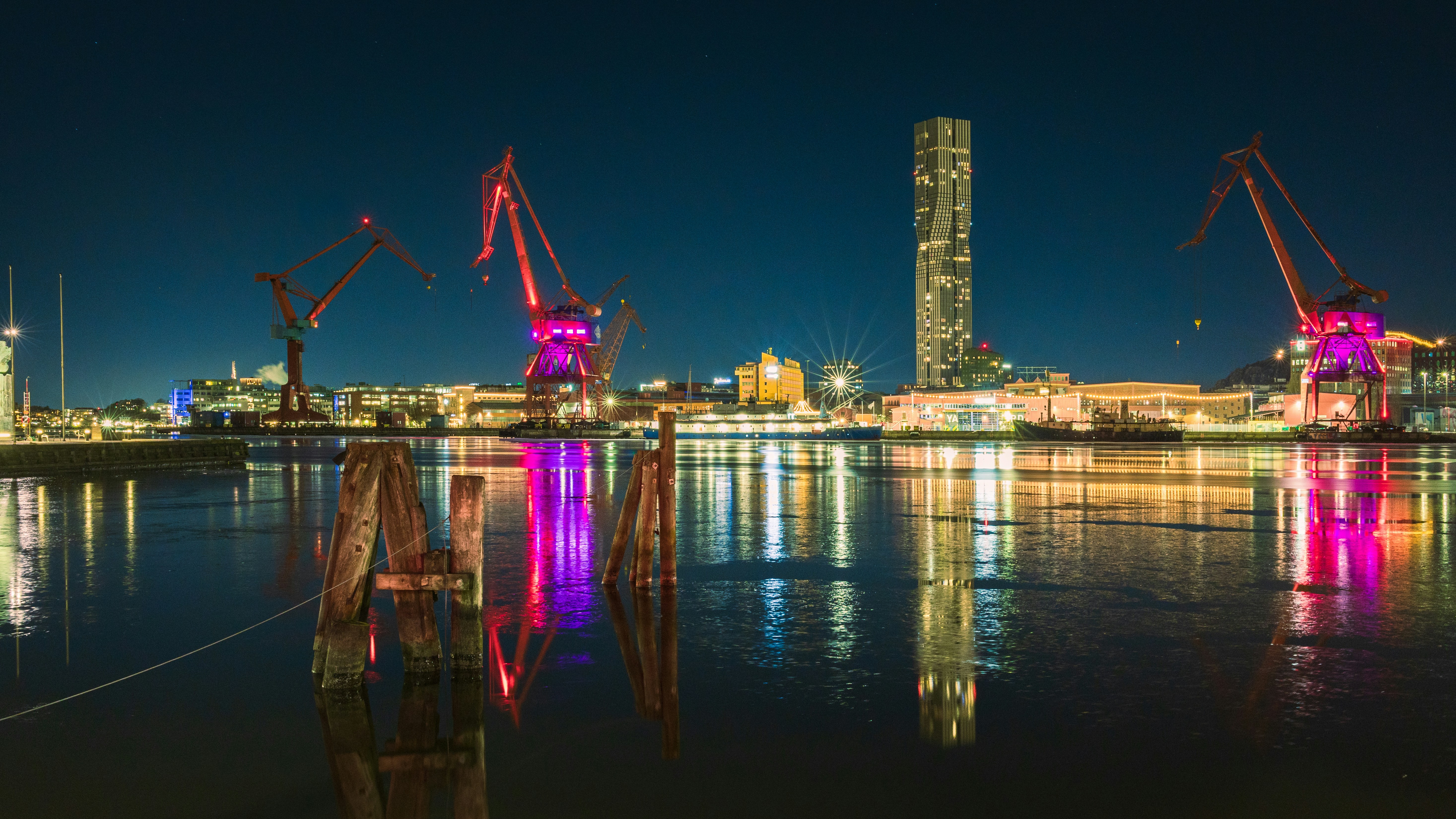 Cranes and buildings illuminated at night by a harbor.