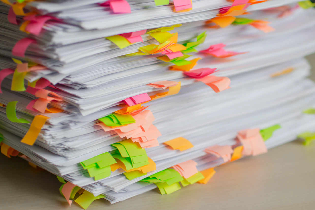 A tall stack of paperwork flagged with sticky tabs, the manual reconciliation a synced system is meant to retire