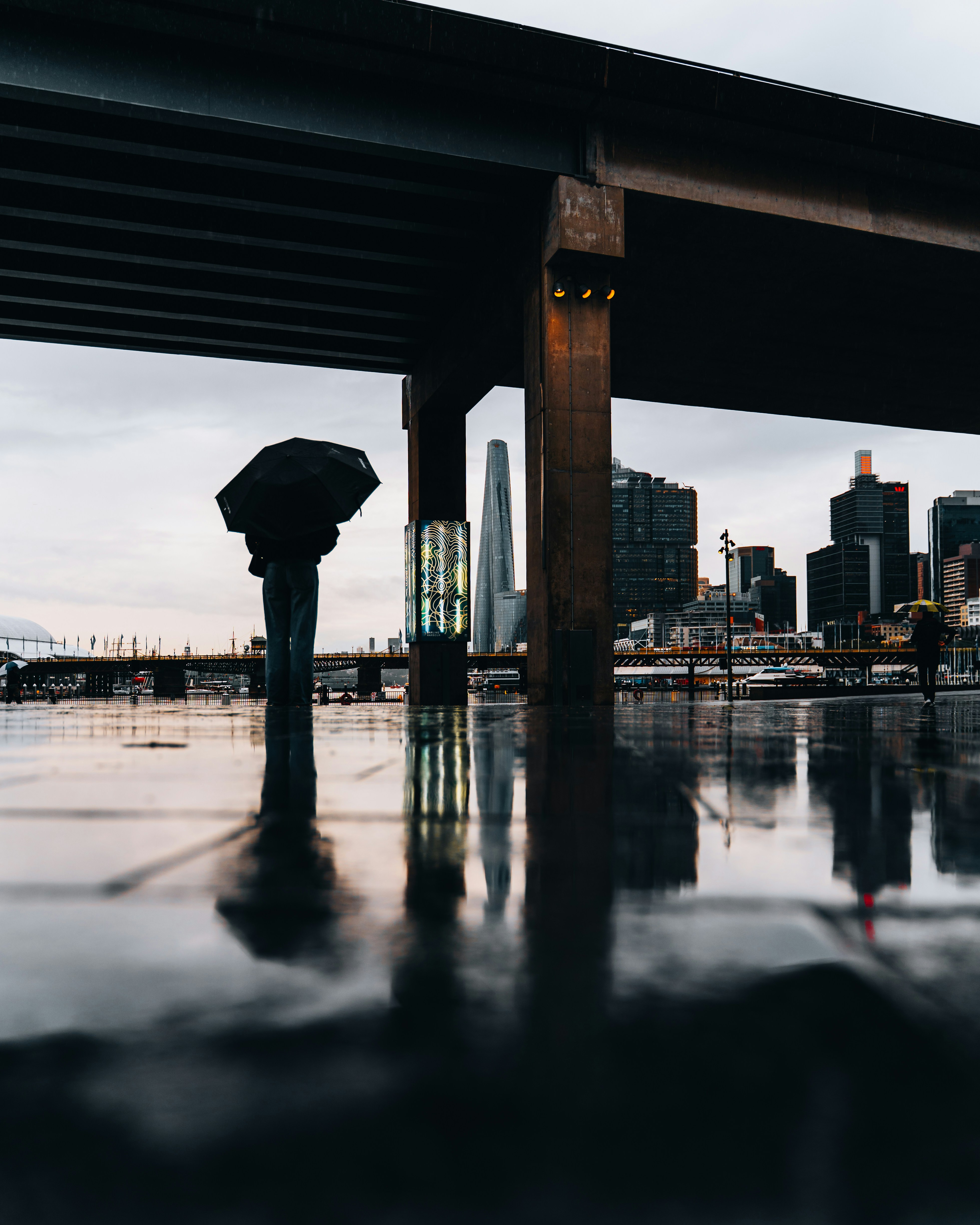 A lone figure with an umbrella under a bridge.