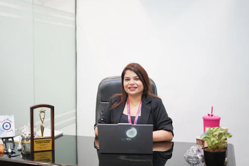 A smiling woman sits at a desk with a laptop