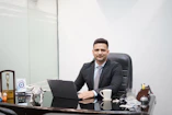 Man in suit sitting at desk with laptop and coffee.