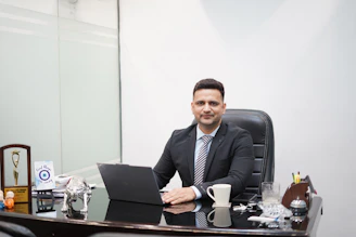Man in suit sitting at desk with laptop and coffee.
