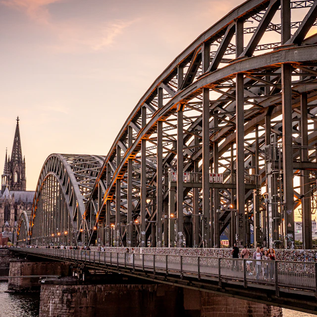 A steel bridge spanning the Rhine with Cologne Cathedral rising behind it in evening light