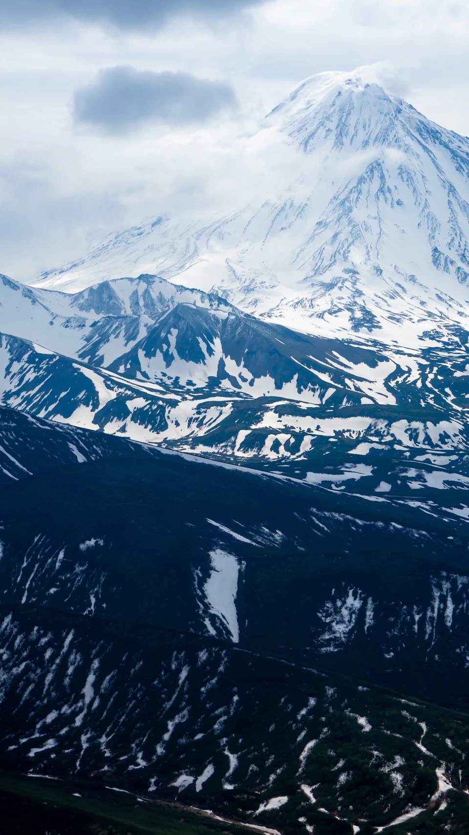 The image presents a breathtaking view of a snow-capped mountain range, with a large peak dominating the scene against an overcast sky. The mountains are blanketed in pristine white snow and exhibit a variety of shades of blue and green, indicating the presence of vegetation at different elevations.

The perspective of the photo is from below, looking up towards the towering mountains. This vantage point provides a sense of scale and grandeur to the majestic peaks, emphasizing their impressive height and beauty. The overcast sky casts a soft light on the snow-covered landscape, enhancing the overall mood and atmosphere of the image.