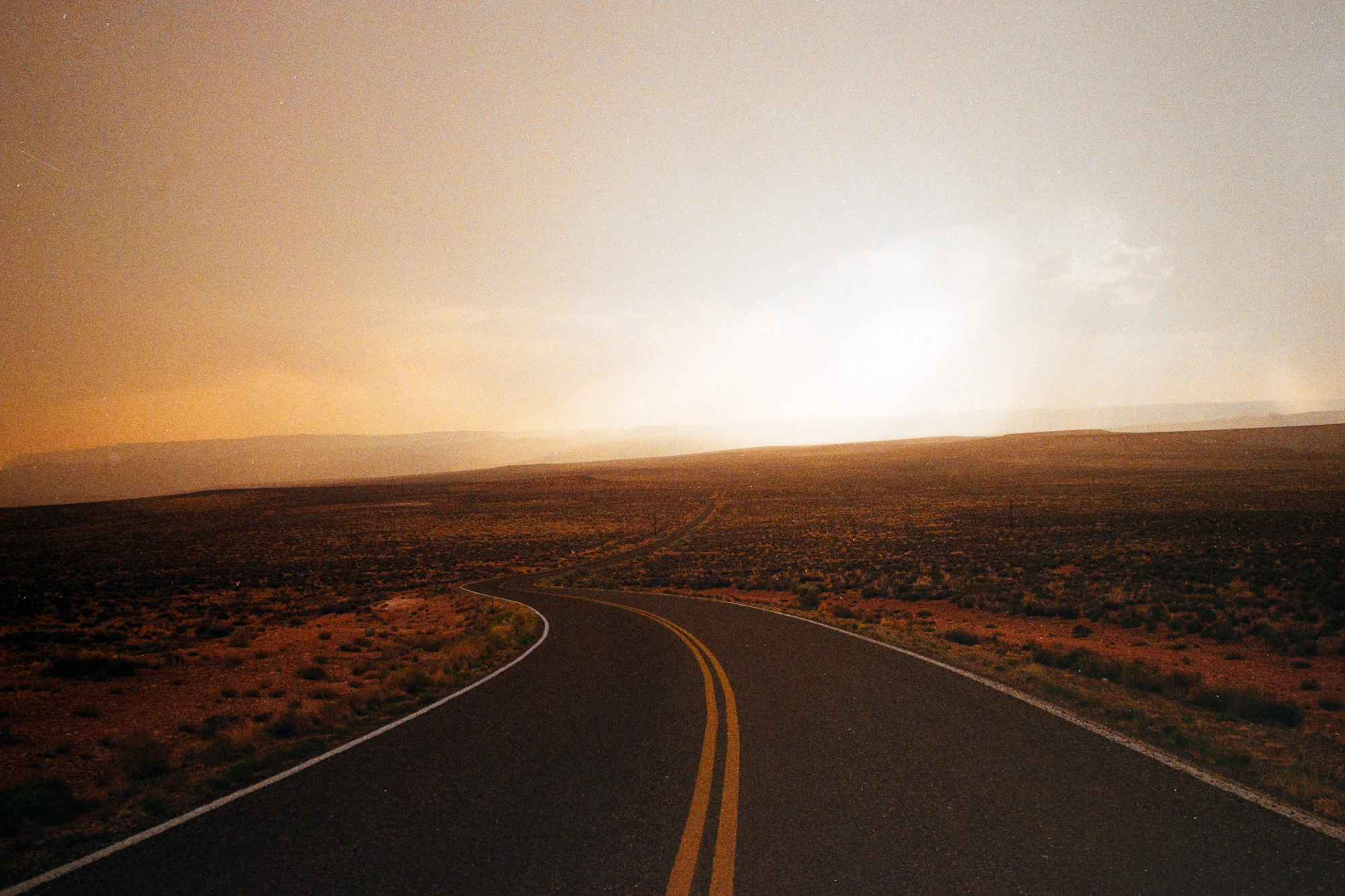 An empty two-lane road curving through open country at dusk, warm haze on the horizon.