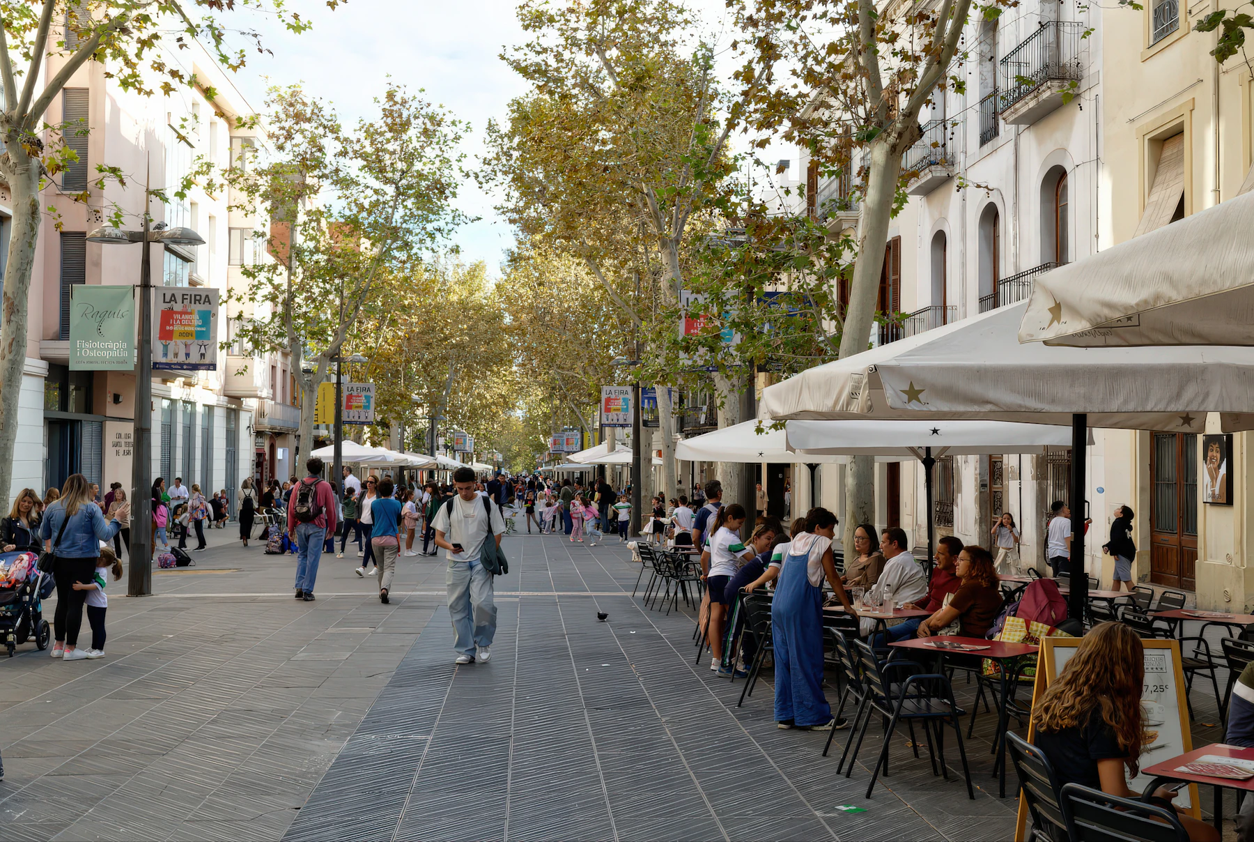 A tree-lined pedestrian street with people walking on the left and a full cafe terrace under white umbrellas on the right