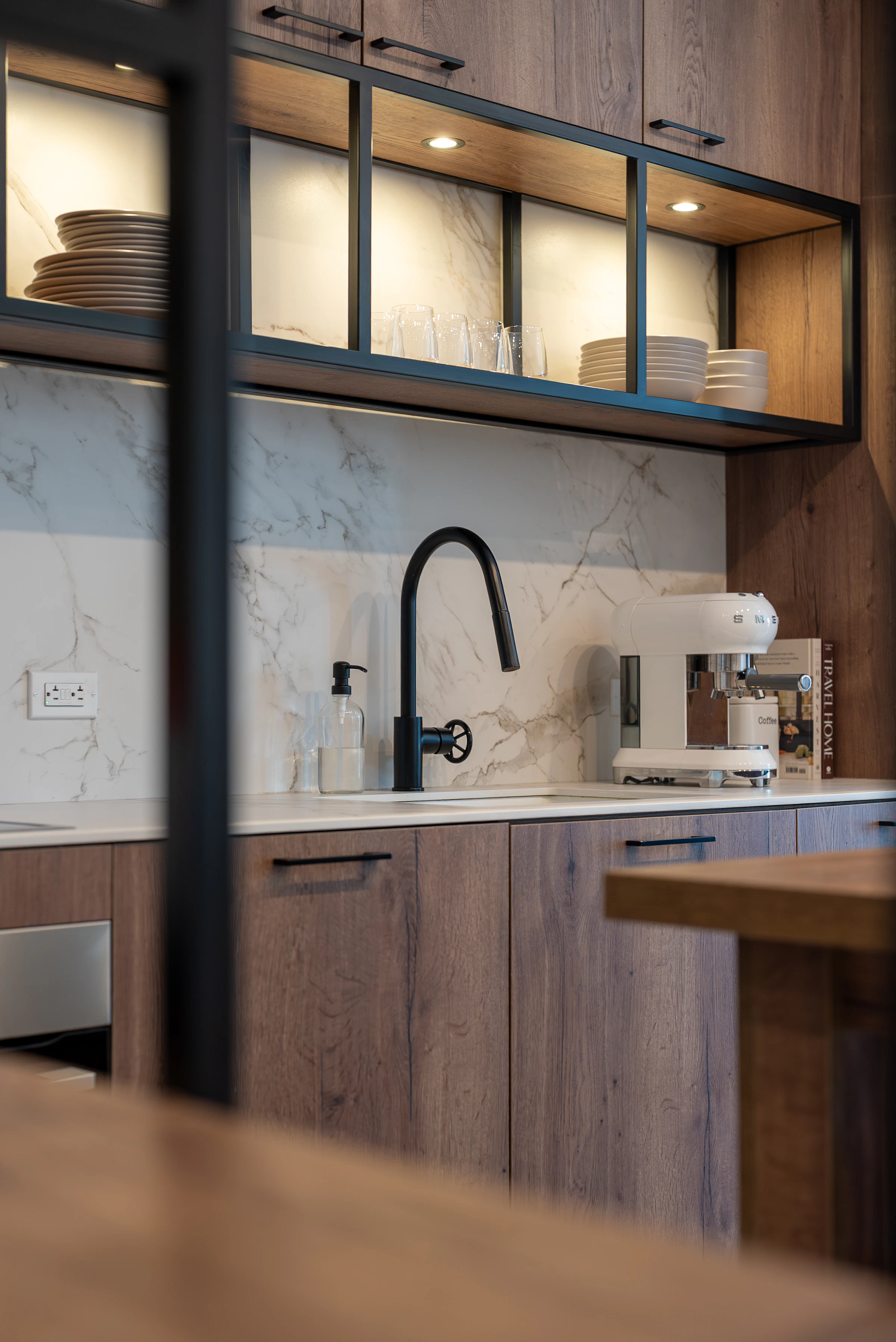 Close-up of a contemporary kitchen with dark wood cabinetry, marble-look backsplash, black-framed glass shelving, a black faucet, sink, and countertop coffee machine.